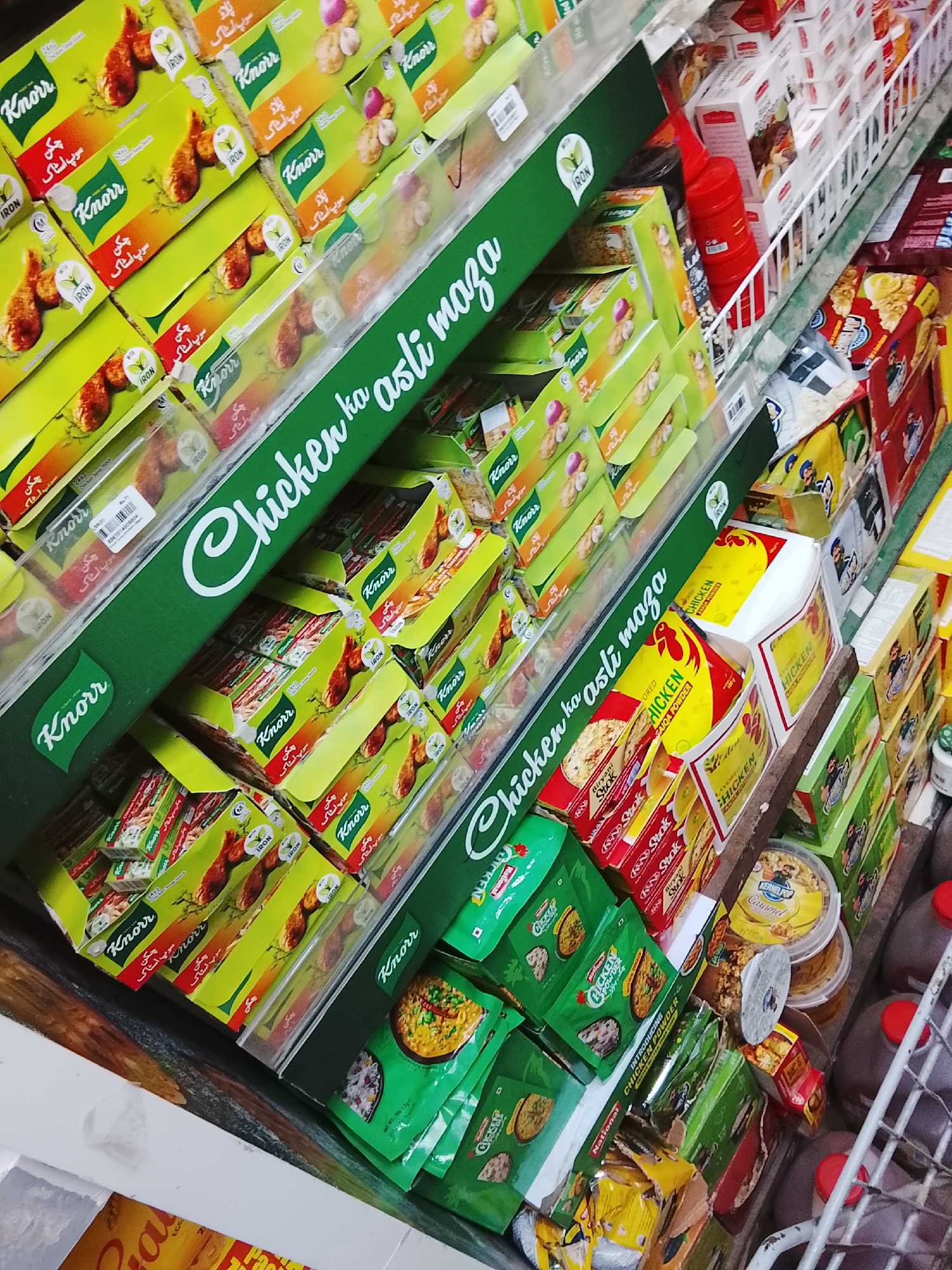The image shows a shelf stocked with various food products, seemingly inside a shop. The most prominent products are from the brand "Knorr," specifically "Chicken Soup Stock" and other chicken-flavored items. Many packages have labels written in Urdu and English. The shelf also contains products from the brand “National” for chicken powder, and “Ken Stok”. The area is likely a grocery store or a small shop in a residential area. Based on the identified location, it is likely within the Gulgasht Colony in Pakistan.