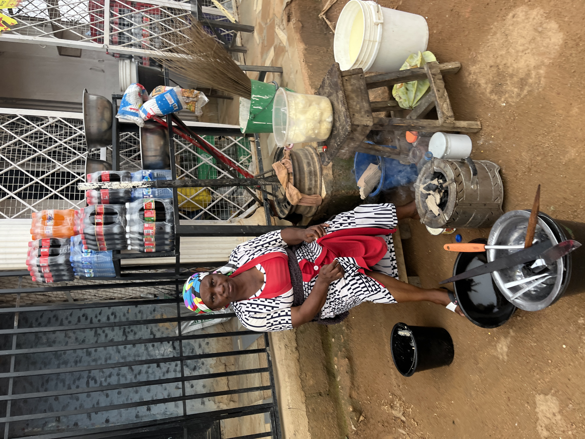 A woman in a black and white patterned dress with a red apron sits on the ground next to a collection of items for sale, likely in a market setting in Jos, Nigeria. She is wearing a colorful headscarf. In front of her are stacks of bottled drinks, including orange soda and water. Behind her, a makeshift cooking station is set up with a small charcoal stove emitting smoke. Various buckets and containers, some filled with food items, are also visible. A broom leans against a shelf. The ground is earthy and appears to be a dirt pathway. The overall scene suggests an outdoor market or street vendor operation. The image is oriented vertically, as if taken while standing and tilting the camera down.