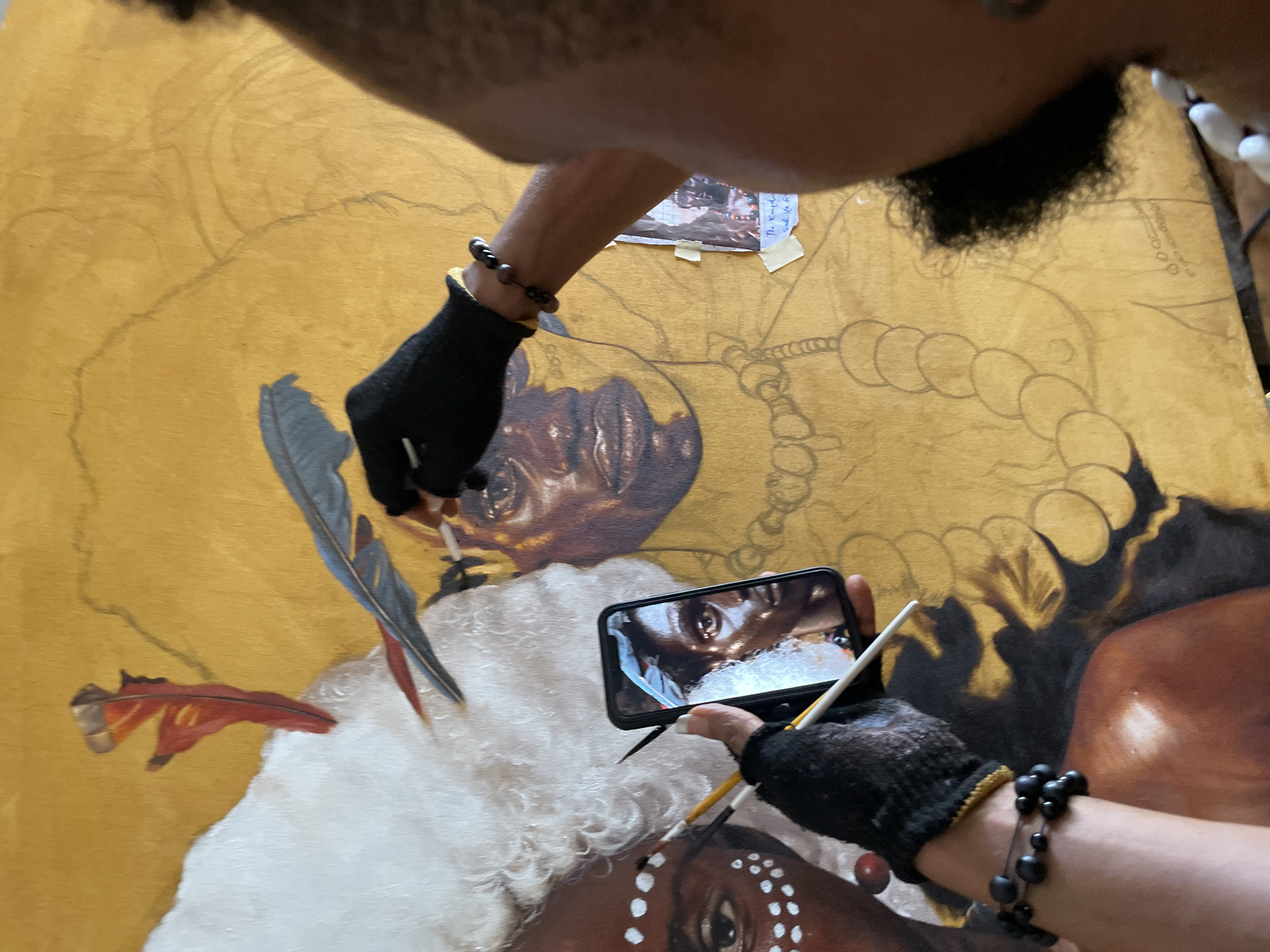 An artist is painting a portrait on a gold-toned canvas. The artist uses a smartphone to view a reference image while working. Two hands wearing black gloves and bead bracelets are visible. One hand holds a paintbrush and the phone, while the other holds a paintbrush near a grey and red feather. The portrait shows an African person with white face paint and a white headdress with feathers. The painting is set on a surface, and the artist's torso is partially visible in the upper area of the frame. The location is presumed to be Ado, Nigeria.