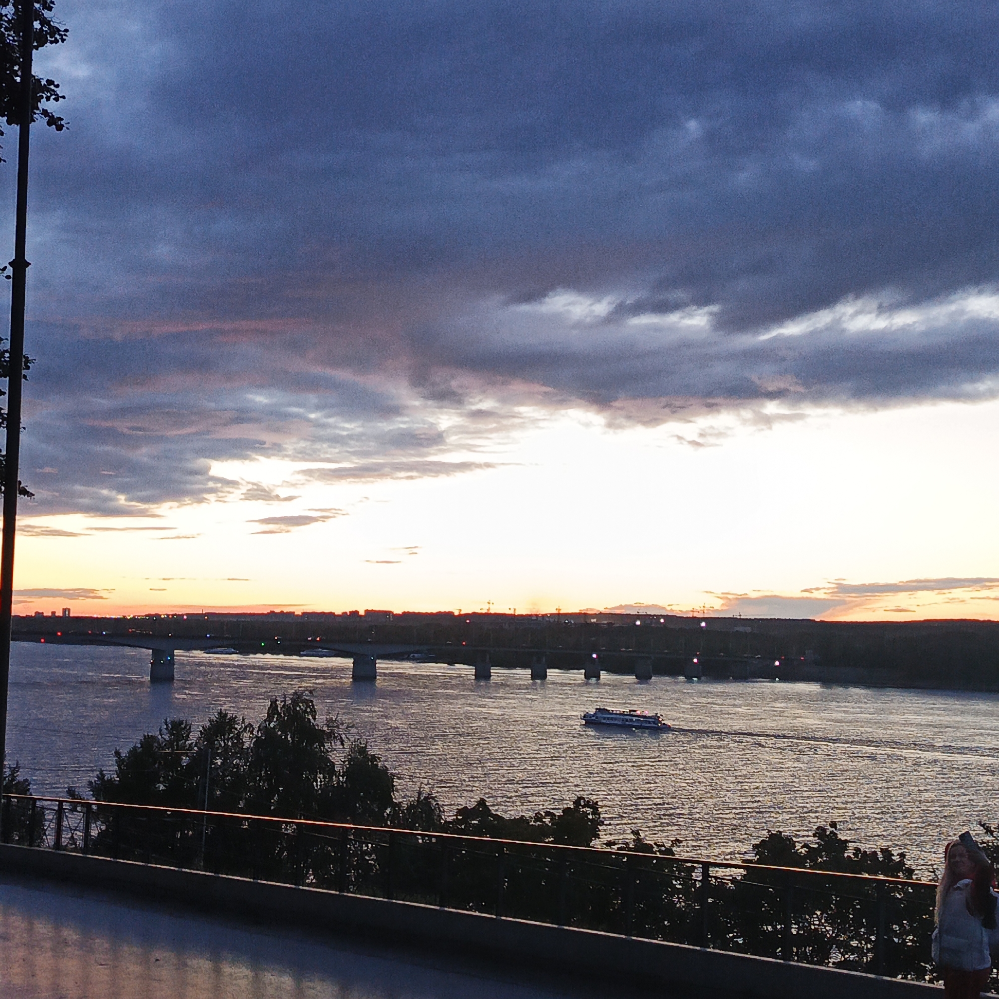 The image depicts a twilight scene over the Kama River in the city of Perm, Russia. In the foreground, a dark balcony railing overlooks the water. Beyond the railing, trees are silhouetted against the sky. A wide river flows towards the horizon, where a bridge spans its width. The bridge is illuminated by scattered lights, suggesting it is a significant infrastructure. A passenger boat is visible on the river, moving from left to right. The sky is filled with dramatic clouds, transitioning from dark blues at the top to soft oranges and pinks near the horizon, indicating either sunrise or sunset. In the far right of the image, a woman with blonde hair is partially visible, looking out at the river. The overall atmosphere is serene and picturesque.