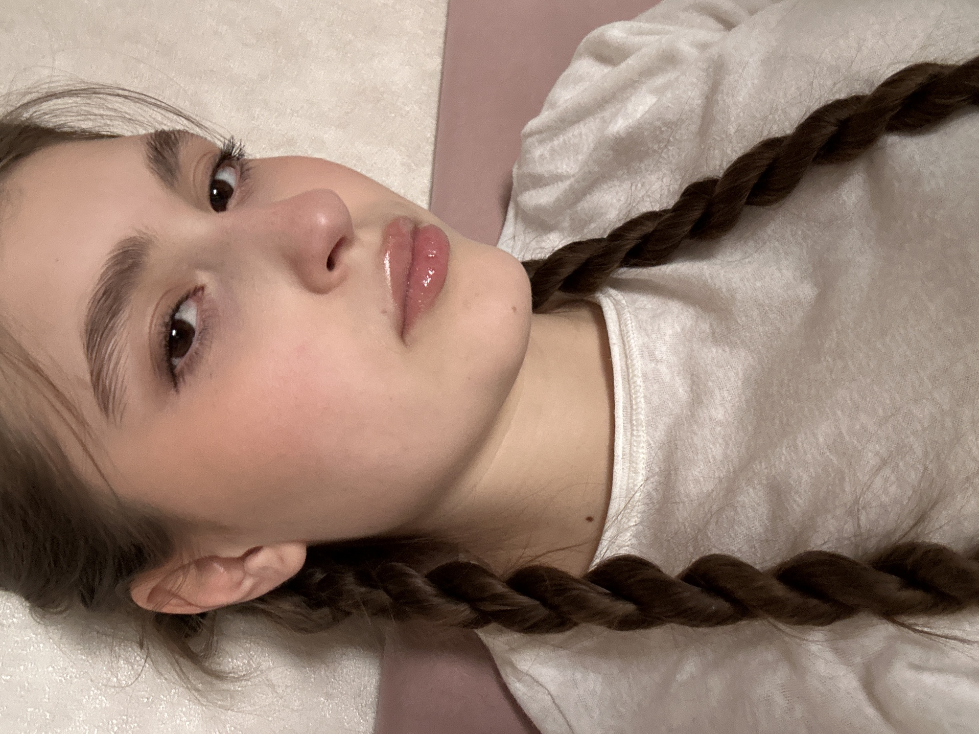 A close-up, high-angle photograph captures a young person, likely female, lying down and looking towards the camera. The subject is positioned diagonally across the frame, with her head in the upper left and her body extending towards the lower right.
Her face is the central focus, showcasing fair skin with a natural, soft appearance. Her eyes are open, with dark irises, and she has long, prominent eyelashes, suggesting mascara. Her eyebrows are full, dark, and well-groomed. A small, slightly upturned nose is visible, and her lips are full and appear to be coated with a glossy, light pink or clear lip product, reflecting light. A subtle blush is visible on her cheeks.
Her dark brown hair is styled in two thick, twisted braids that run alongside her body, one visible on the upper right and the other across her lower body. The texture of the braids is clear, showing the individual strands twisted together. She is wearing a light-colored, possibly white or off-white, plain t-shirt or top with a slightly ribbed texture visible around the shoulder/collar area. A small, dark mole is visible on her neck or upper shoulder area, near the collar of her shirt.
The background consists of two distinct areas: a light beige or off-white surface that covers most of the left side and top, and a solid light pink or muted purple strip running diagonally behind her, parallel to her body. This suggests she is lying on a bed or a surface with a colored panel.
The lighting is soft and even, highlighting the gloss on her lips and the contours of her face, indicating an indoor setting. Her expression is calm and neutral, as if posing for the photograph. No text is visible in the image.