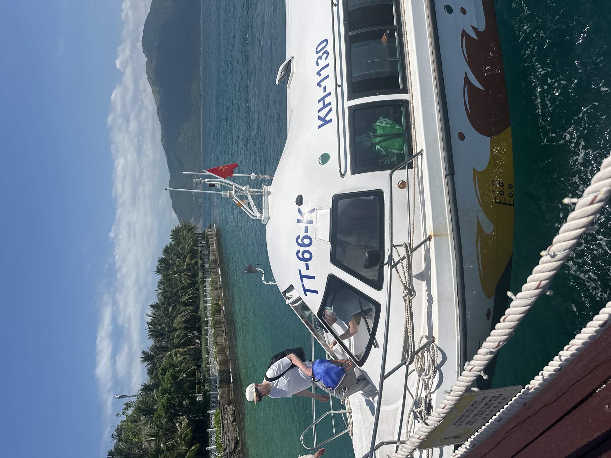 The image captures a sunny coastal scene at a pier in Nha Trang Ward, Vietnam. A white motorboat, marked with registration numbers "TT-66-K" on the front cabin and "KH-1130" on the side, is moored alongside a dark brown wooden pier. The boat's hull features a yellow and brown wave-patterned decal, with draft markings of "1.0", "0.8", and "0.6" visible on its side. A red Vietnamese flag with a yellow star flies from an antenna mast atop the boat's cabin.
A man wearing a white baseball cap, white t-shirt, and light grey trousers, carrying a black backpack and a blue drawstring bag, is stepping from the boat onto the pier. The arm of another individual is partially visible to the left, near the boat's boarding area. Through the boat's tinted windows, at least two persons are visible inside the cabin, one wearing a green life vest.
The wooden pier has a white rope railing secured by white posts. Attached to this railing is a yellow warning sign, partially obscured, which reads "PLEASE DO NOT LEAN ON THE ROPE" followed by the Vietnamese translation "VUI LÒNG KHÔNG DỰA VÀO DÂY CỘT". The water around the boat and pier is a clear, calm turquoise.
The background reveals a lush coastline densely populated with palm trees, behind which a tall, thin antenna tower rises. A white decorative fence or railing borders the shoreline, separating it from the water. Distant green mountains are visible under a bright blue sky with scattered white clouds.