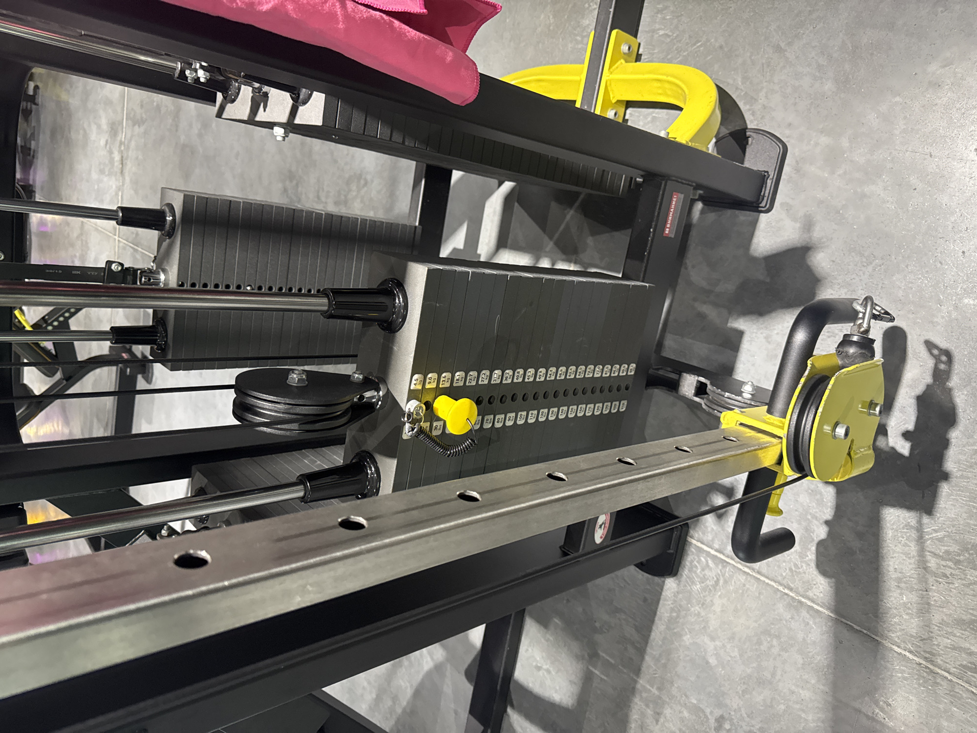 This is a medium-shot, eye-level, indoor photograph of a cable crossover machine in a gym. The machine is black with yellow accents and features a stack of weight plates. A pink towel is draped over the side of the machine. The floor is concrete. The gym is located in Komsomolsk-on-Amur, Russia.