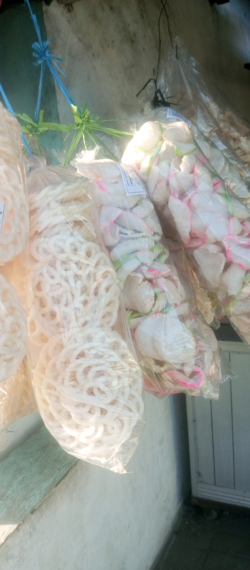 The image shows multiple plastic bags of crackers, likely kerupuk, hanging outdoors. In the foreground, to the left, is a bag filled with white, ring-shaped crackers. To the right of this bag, and further into the background, are bags containing irregularly shaped, flat crackers that are white with pink and green accents. The crackers appear to be commercially packaged, as one bag shows a label with text. They are suspended by blue and green ties made of what appears to be plastic string or natural fibers. The background consists of a textured, light-colored wall and a sliver of a gray door with vertical panels. The lighting suggests it is daytime, with sunlight casting shadows. The scene is consistent with a street-side vendor or a shop stall in Kediri City, Indonesia, where such snacks are commonly sold.