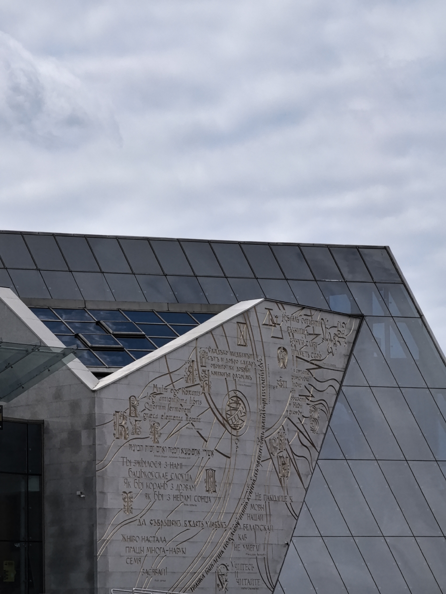 The image shows the facade of a modern building with a glass roof and a concrete wall featuring an inscription in multiple languages. The inscription is a relief artwork with text carved into the concrete surface. Some of the text is in Latin, and some appears to be in Cyrillic script. The building is angular and has a contemporary architectural style. The sky above is overcast with visible clouds. There are no people or animals visible in the image. The scene appears to be an outdoor architectural shot focusing on the building's exterior details.