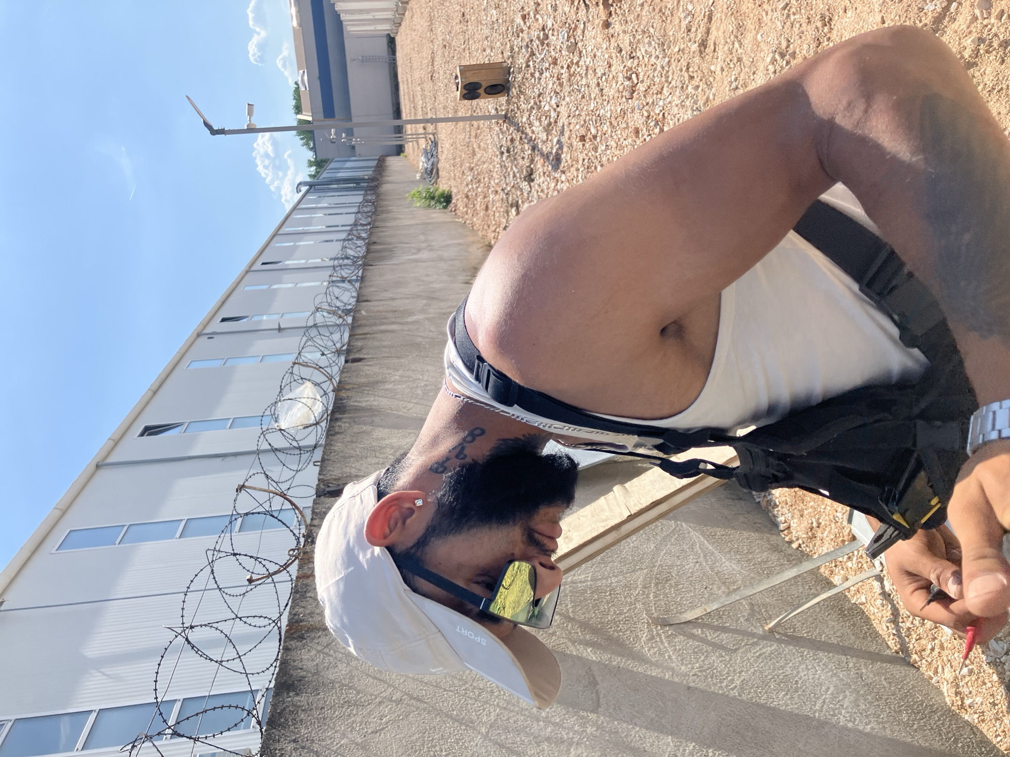 A photograph taken in Plovdiv, Bulgaria, shows a man wearing a white tank top and a black harness or vest, standing near a building with barbed wire along the top of the wall. He is also wearing a light-colored cap with the word "SPORT" on it and mirrored sunglasses. The building is long and appears to have multiple windows. In the background, there is a stretch of land with gravel and some vegetation, along with a light pole and a building further away. The sky is clear and blue. The man is holding a small red object in his hand, possibly a tool or wire. The scene suggests an industrial or semi-industrial location. The photo is taken from a slightly tilted angle.