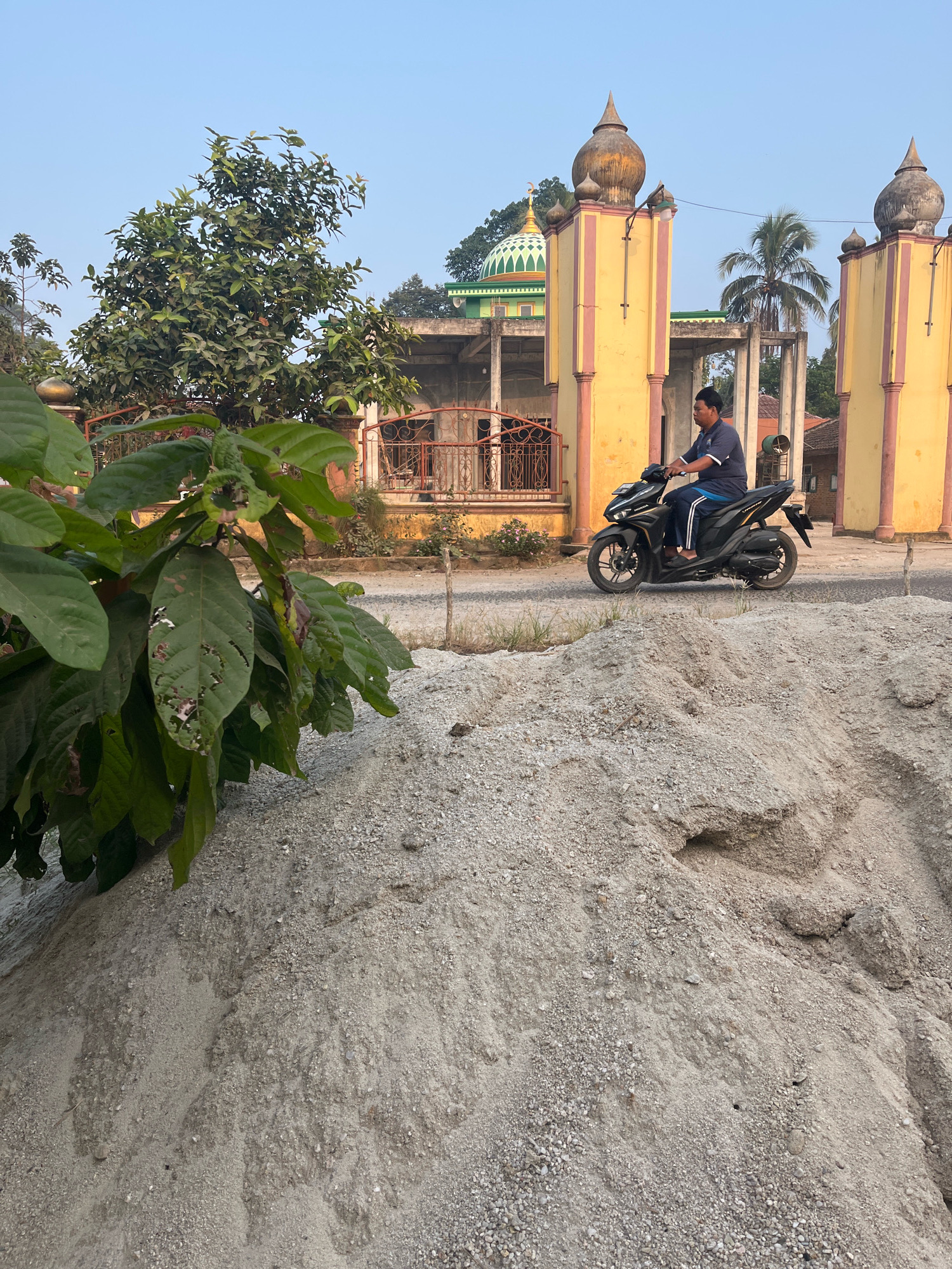 A man rides a black scooter on a paved road in front of a mosque with yellow pillars and domes in Kebumen, Indonesia. In the foreground, a pile of sand is visible. The mosque has a green dome and is surrounded by trees and a fence. The sky is clear and blue.