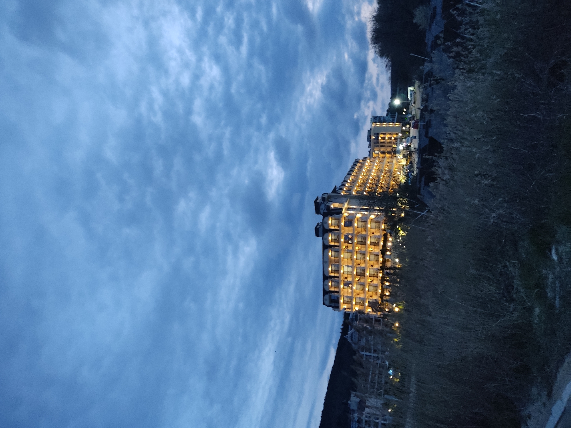 The image shows a multi-story building with illuminated windows, likely a hotel or apartment complex, under a cloudy evening sky. The building is situated near vegetation, possibly overlooking a coastline. In the lower part of the image, some other buildings are faintly visible. The location is in the city of Unknown, Unknown.