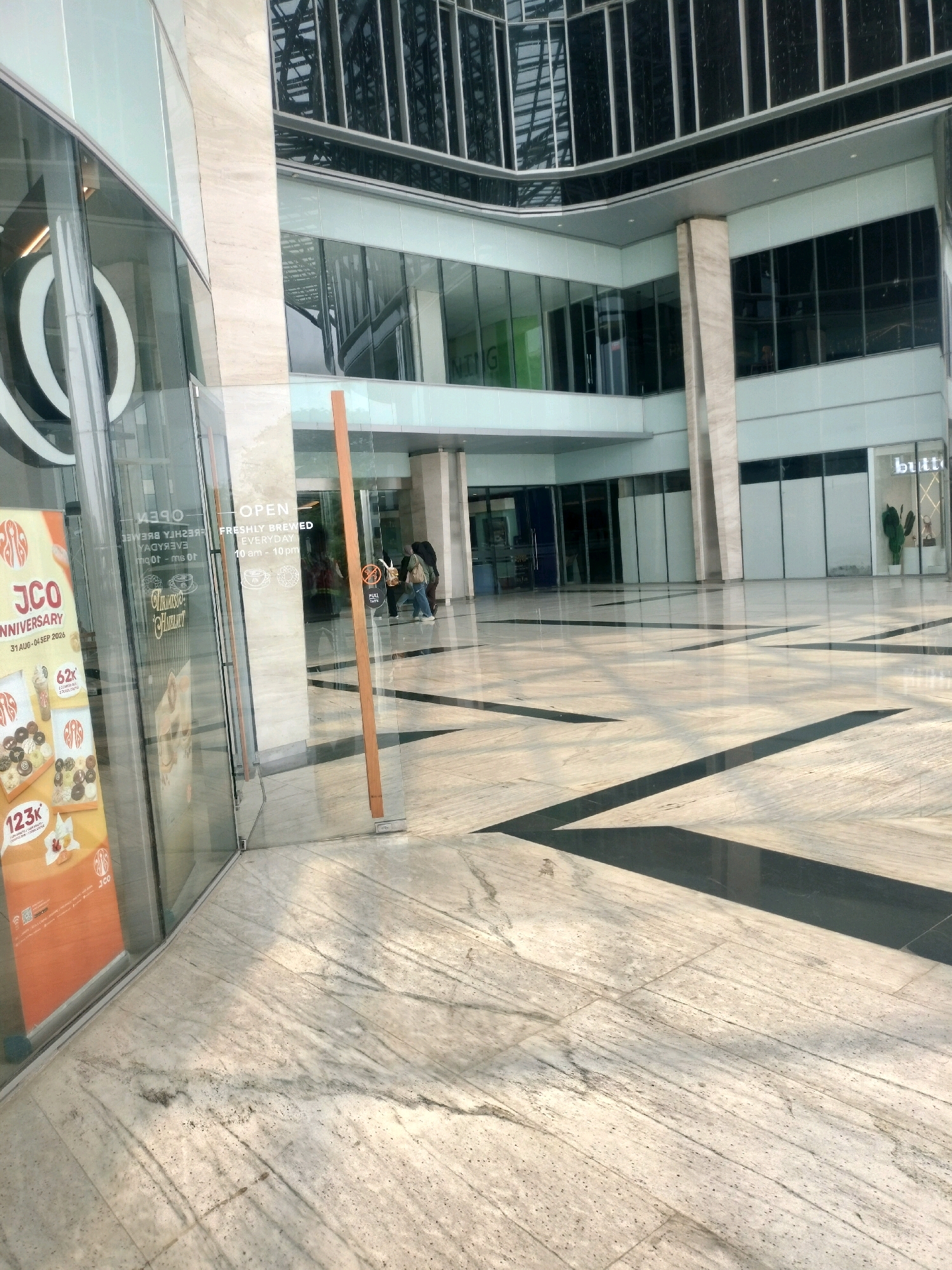 This image captures an exterior view of a modern commercial building in Batam, Indonesia. The architecture features a combination of glass facades and marble cladding. In the foreground, a large glass door leads into what appears to be a retail space. A promotional banner for "JCO Anniversary" is prominently displayed to the left of the entrance, advertising donuts and a special offer. The floor is paved with marble tiles, with dark geometric inlays creating a visually striking pattern that reflects the overhead lighting. The exterior walls of the building are made of reflective glass and light-colored stone. In the background, there are multiple levels of windows, suggesting a multi-story structure. The sky is not visible, indicating this is likely taken in an open-air or covered walkway area within the complex. The scene is well-lit, with sunlight casting shadows across the floor. Two individuals are seen walking away from the camera towards the building's entrance.