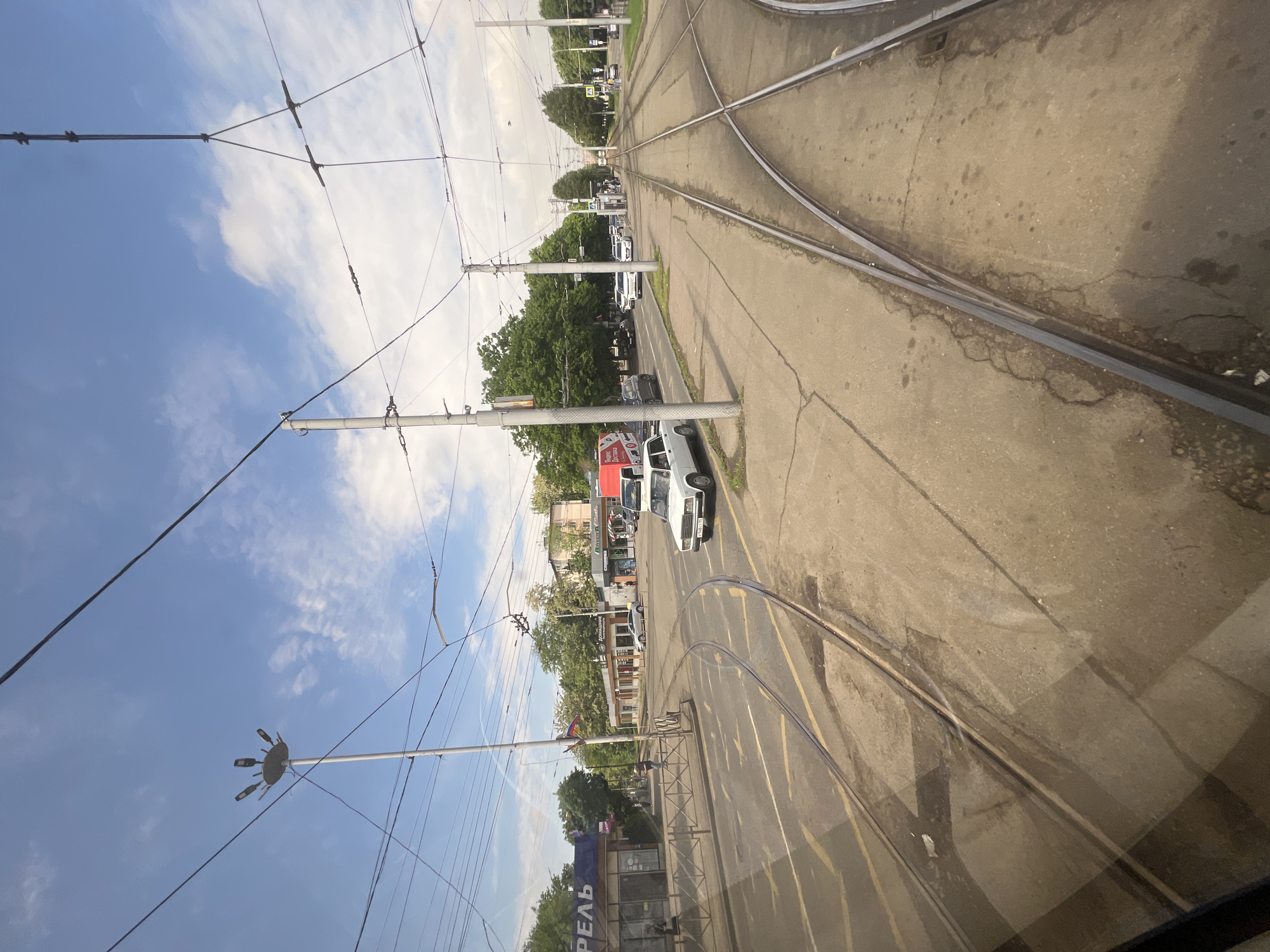The image depicts a street scene in Krasnodar, Russia, featuring tram tracks in the foreground. The view is taken from inside a vehicle, likely a tram or a bus. The tram tracks intersect, suggesting a junction. Overhead power lines and poles are visible, supporting the electrical system for the tram. A line of parked and moving cars are present alongside the street. Buildings and trees line the road, suggesting a residential or commercial area. The sky is blue with some clouds. A sign with the word "РЕАЛЬ" is partially visible in the lower left corner.