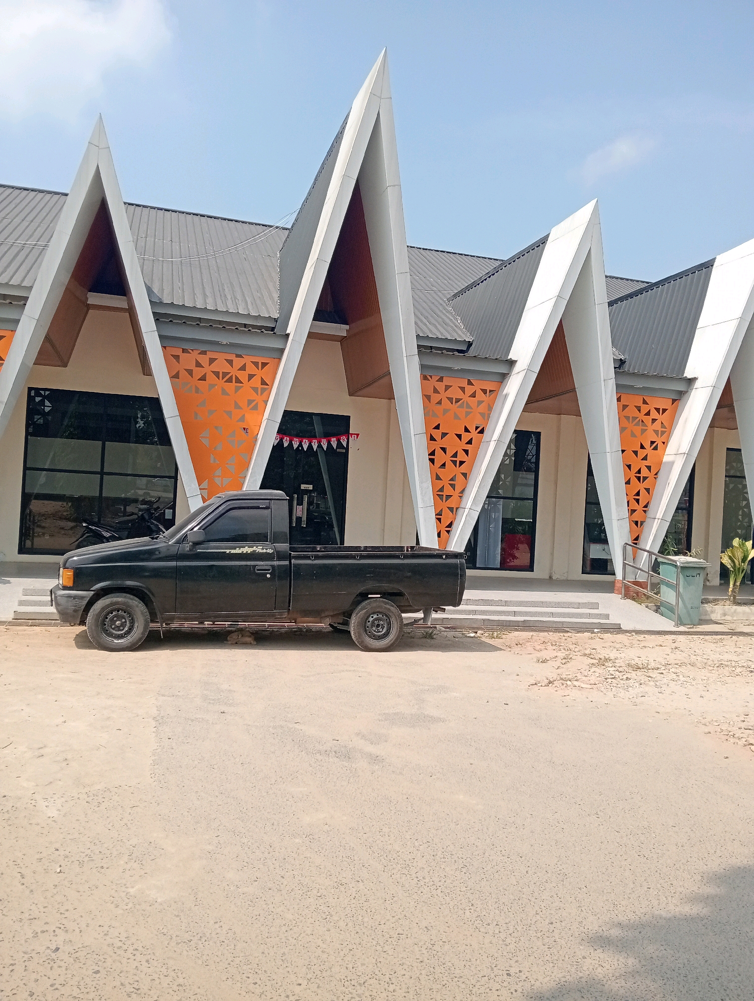 A black pickup truck is parked on a dirt road in front of a building with a distinctive modern roofline. The building has a light-colored facade, large dark windows, and decorative orange panels with triangular cutouts. The roof is made of gray corrugated metal and features several pointed, white architectural elements. A ramp with a metal handrail leads to the entrance of the building, next to which is a green trash bin. A string of red and white pennants is visible above one of the entrances. The sky is clear blue with a few wispy clouds. The scene appears to be in Pringsewu, Indonesia, based on the context provided.