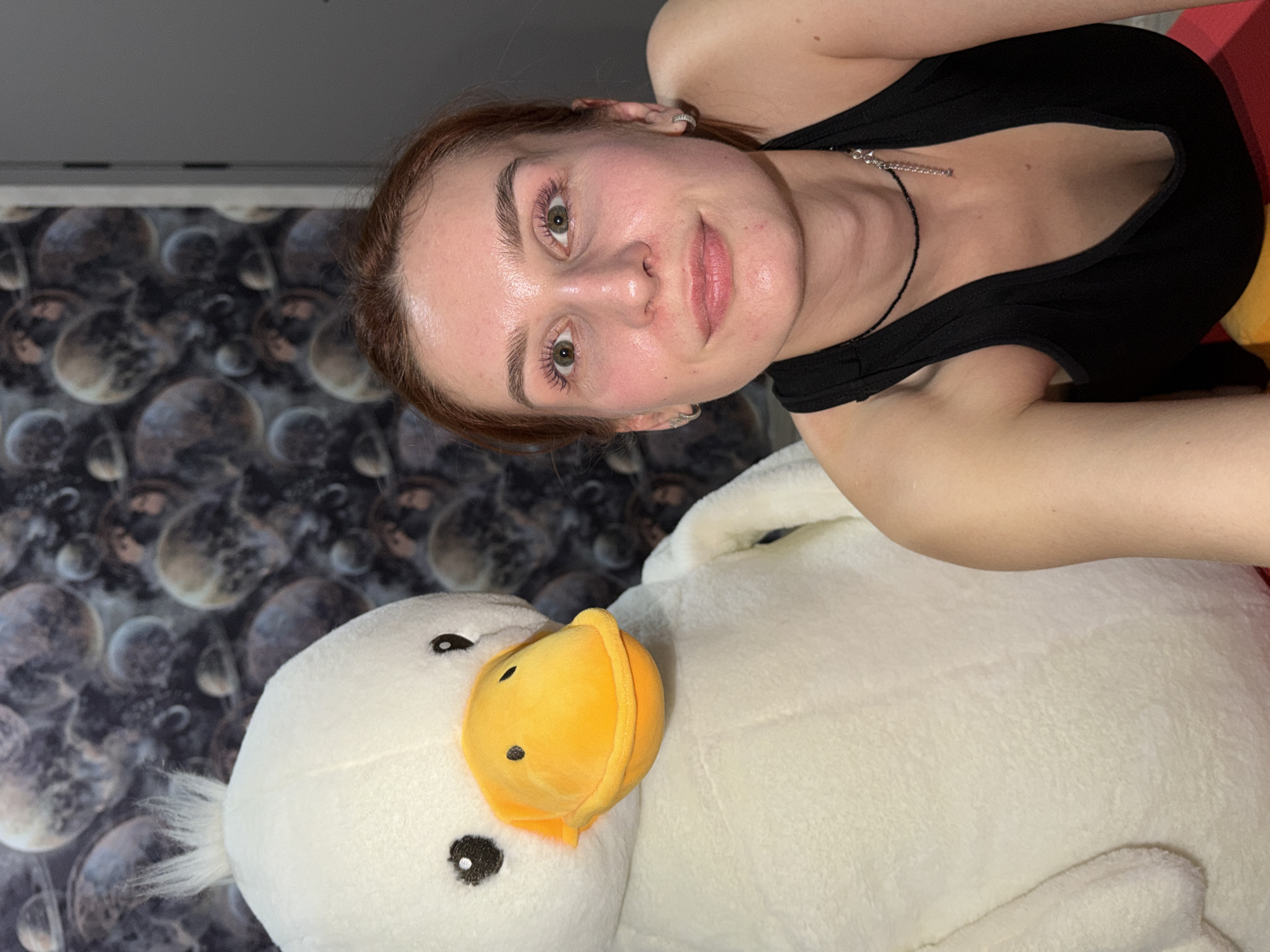 A selfie-style photo shows a young woman with auburn hair, fair skin, and green eyes, wearing a black tank top and a delicate necklace, lying on a large white plush duck toy. The background features a patterned wallpaper resembling planets or celestial bodies. The photo was likely taken indoors in an unknown location, possibly in the city of Chaikovsky, Russia.