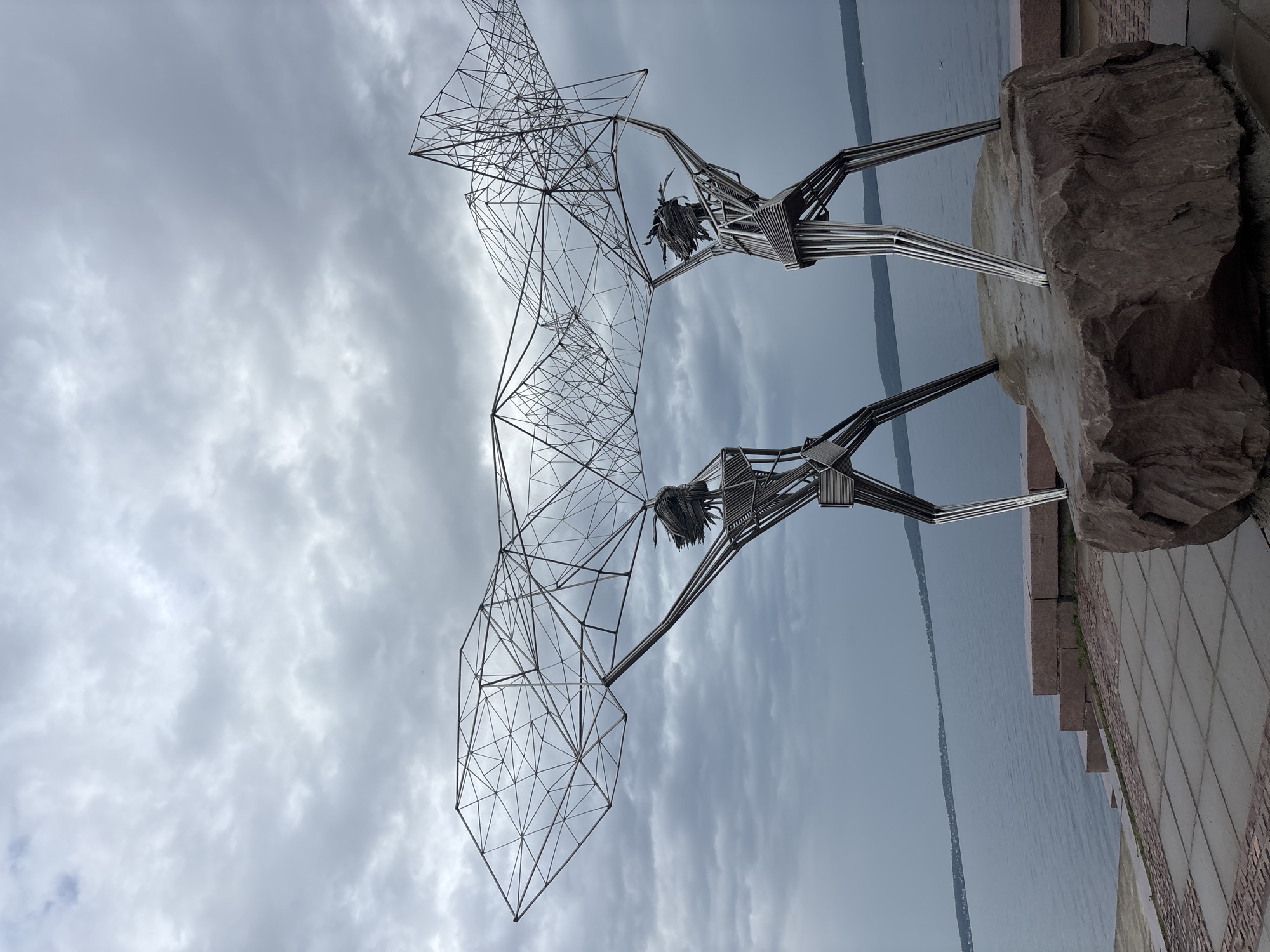 This media file features an outdoor sculpture depicting two stylized figures made of metal. The figures appear to be holding up a large, geometric, cage-like structure composed of interconnected wires. The sculpture is mounted on a large rock and is situated next to a body of water, likely a lake or river, with a distant shoreline visible. The ground in front of the sculpture is paved with square tiles. The sky is overcast with dramatic, cloudy formations. The overall impression is one of a public art installation in a scenic waterfront location. The sculpture's abstract design and the cloudy weather contribute to a somewhat somber or contemplative mood. The time of day appears to be daytime, but the heavy cloud cover suggests it could be overcast or nearing dusk. There is no visible text in the image. The location is identified as Petrozavodsk, Russia.