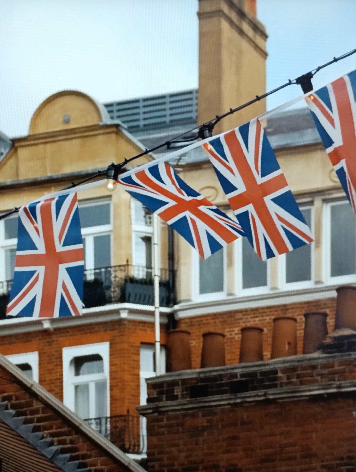 The image depicts a string of Union Jack flags draped across the facade of a building. The flags are triangular and appear to be made of fabric, with the characteristic red, white, and blue pattern of the British flag. They are suspended from a black wire, with several black clips holding them in place. Some of the flags are partially obscured by other flags or by the building's architecture.
The building itself is constructed of red brick and has several windows, some of which are visible behind the flags. The windows have white frames and appear to be closed. There is a balcony with a black railing visible on the second floor. Above the brickwork, there are multiple chimney pots lining the roofline. The sky is overcast and a pale grey.
The scene is set in an urban environment, with the architecture suggesting it could be a residential or commercial building. The presence of the Union Jack flags indicates a possible celebration or patriotic event. The location is identified as the town council of Dubăsari, Moldova.