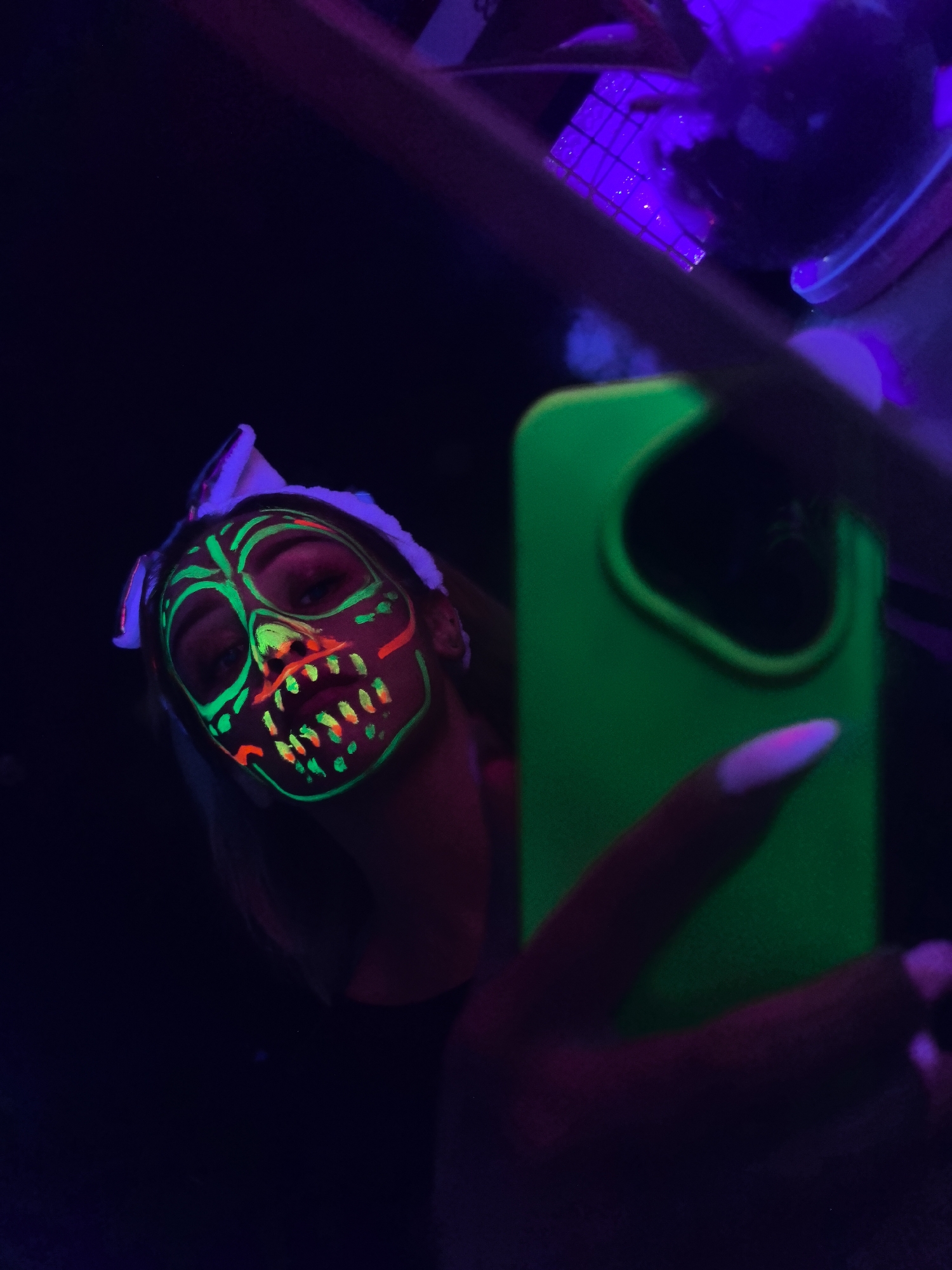 A person is taking a selfie in low light. The person has green and red face paint resembling a monster. They are holding a green phone. The background is dark with some purple lighting. It is not possible to determine the location.