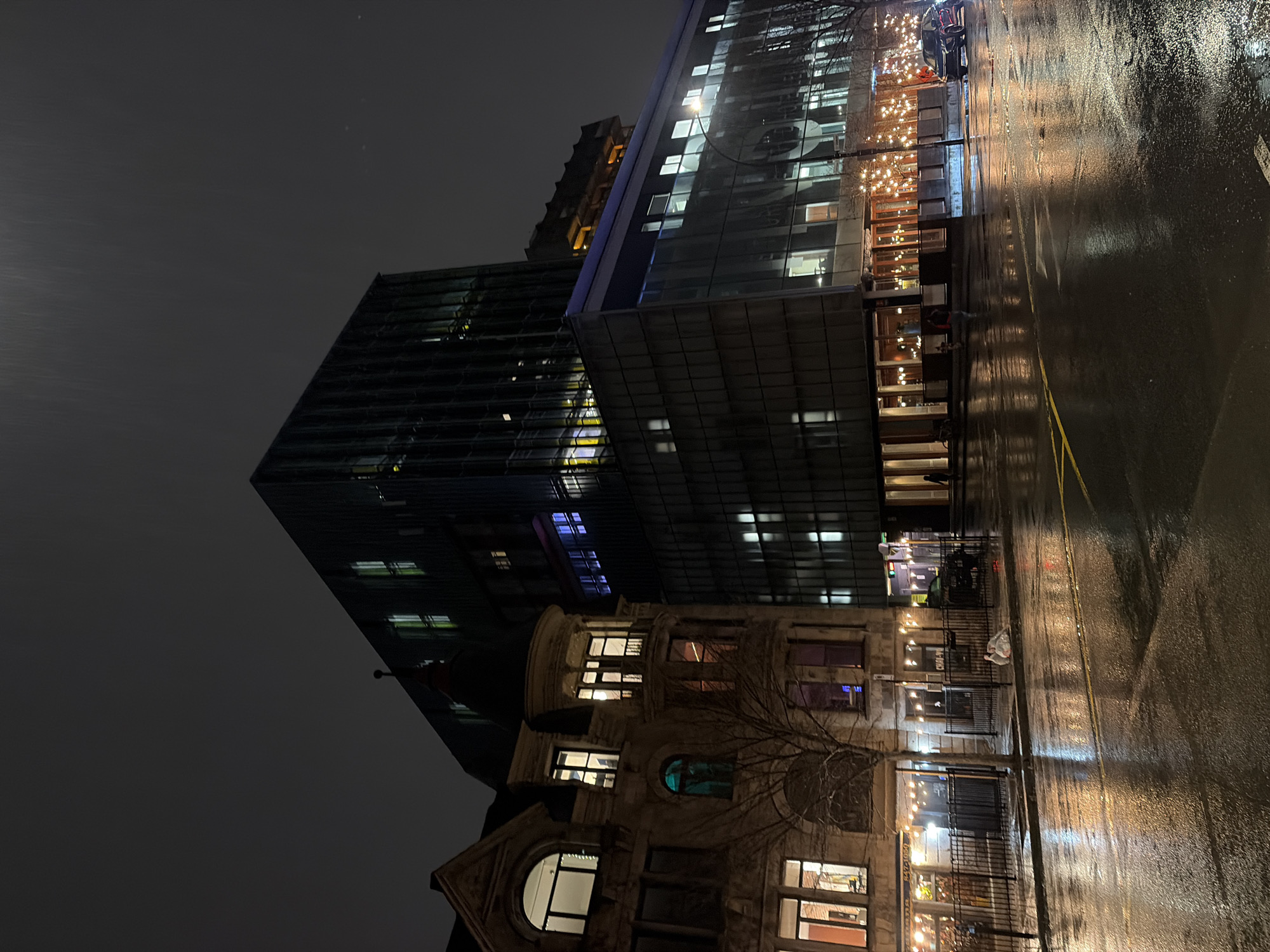 The photograph depicts a street scene at night in Montreal, Canada. The street is wet, reflecting the lights from the buildings. Modern buildings and older brick buildings are present. The modern buildings are taller and have numerous lit windows. Strings of lights are visible hanging outside. A yellow painted line is on the wet pavement. The sky is dark and overcast. A person stands outside of a building.