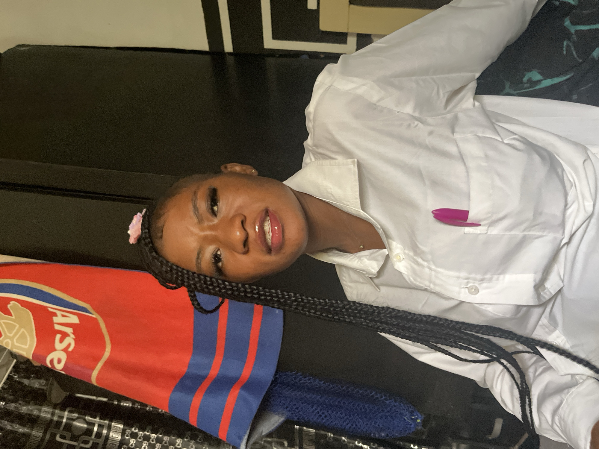 In this media file, a young Black woman is lying on her back, with her head tilted back and her gaze directed upwards. She is wearing a white collared shirt. A pink pen is tucked into the pocket of her shirt. Her hair is styled in long braids, and a small pink hair accessory is visible near her temple.
To her left, an Arsenal Football Club flag is draped. The flag is red with blue and gold accents, and the word "Arse" is visible on it. Beneath the flag, a blue mesh bag is partially visible. The background appears to be a dark surface, possibly a table or a floor, with some geometric patterns.
The lighting suggests an indoor setting, possibly in the evening or under artificial light, as there are no clear indications of natural daylight or weather conditions. The woman's expression is difficult to ascertain precisely due to the angle and crop of the image, but her mouth is slightly open, and her eyes are wide open. The overall scene appears casual and relaxed.
The provided location context is Jos, Nigeria.