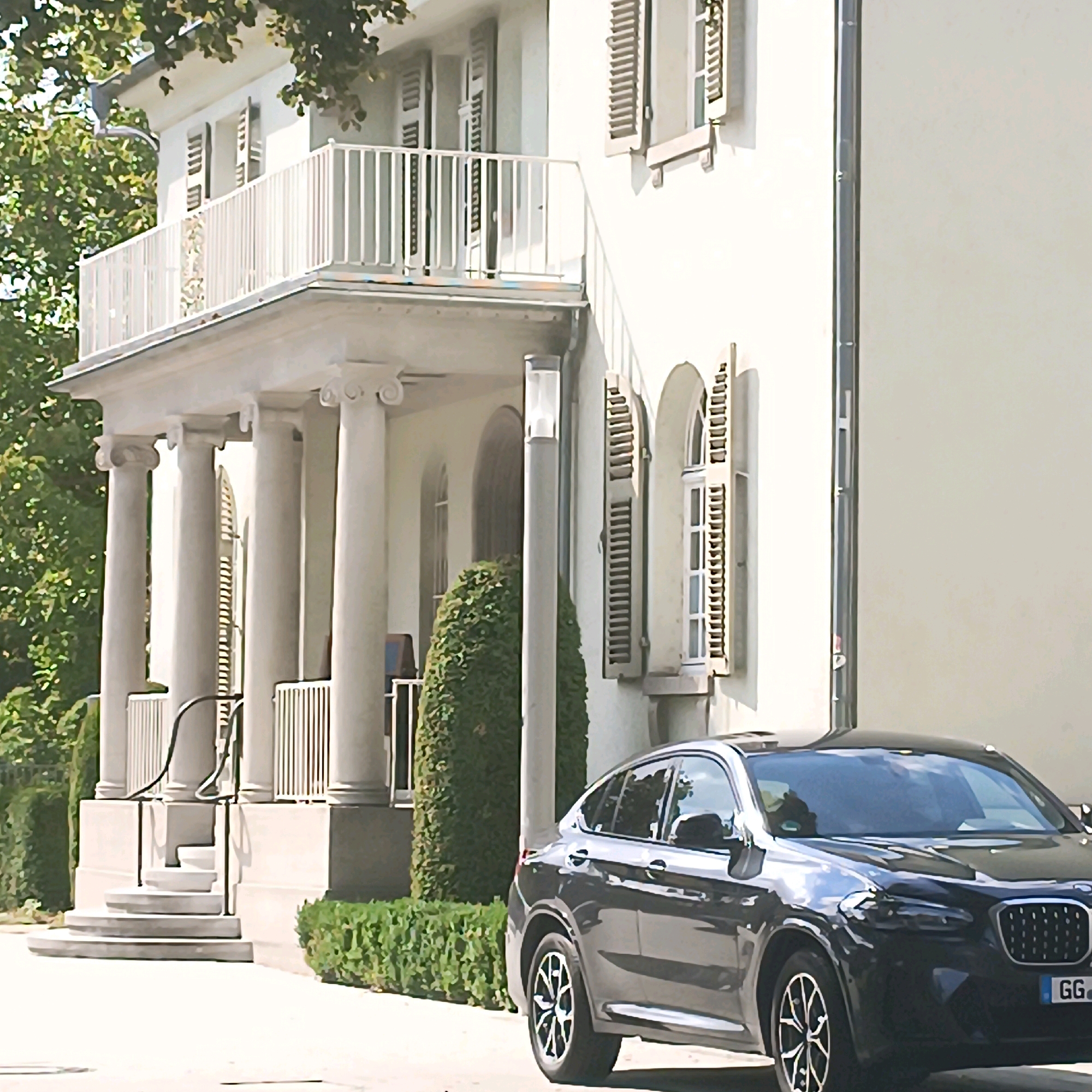 A dark gray BMW X4 is parked in front of a white, multi-story building with classical architectural elements. The building features columns, arched windows with shutters, and a balcony with a white railing. Lush green trees and manicured hedges surround the property. The car's license plate is partially visible, reading "GG". The scene appears to be in a residential or institutional area.