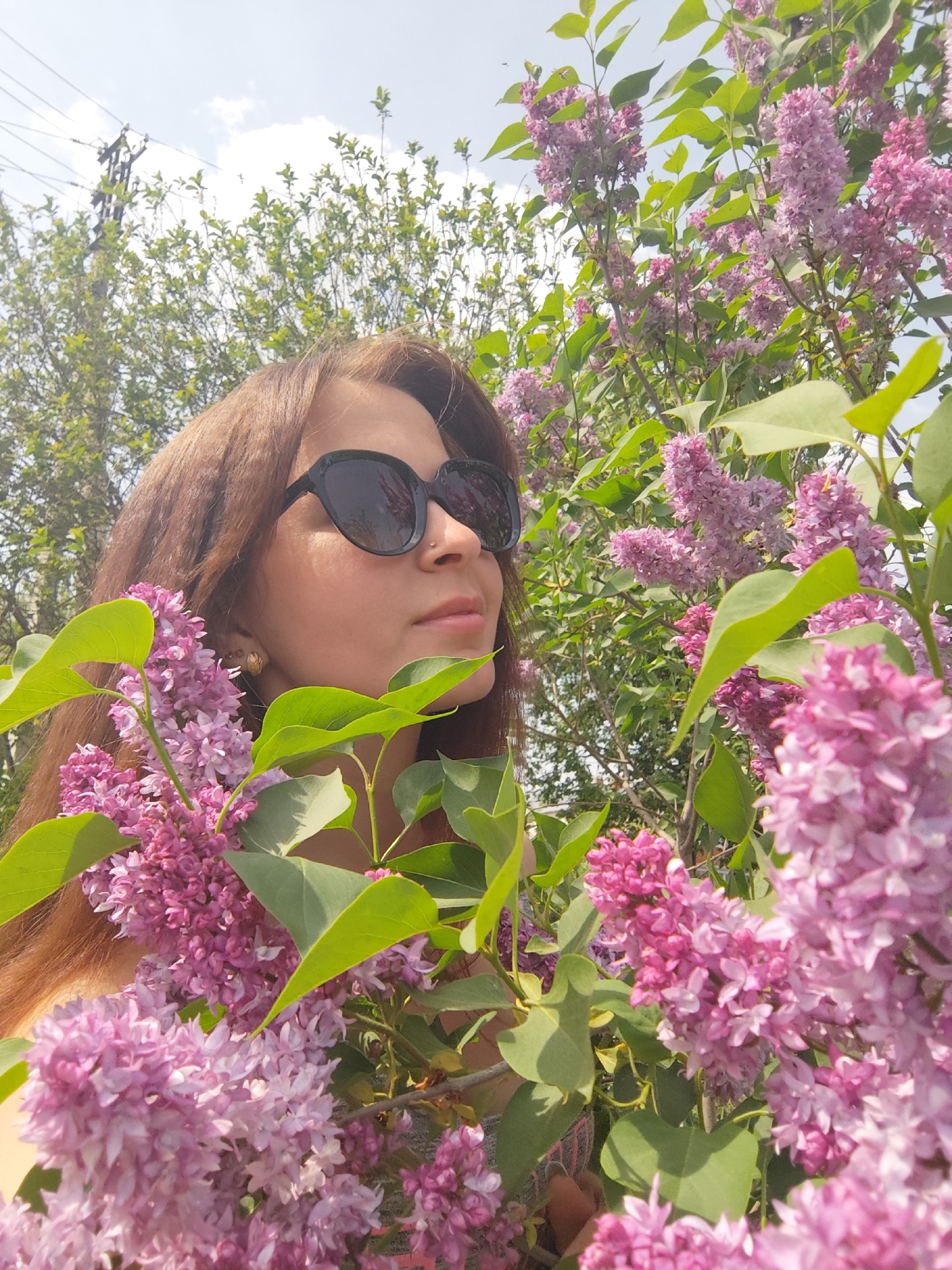 A woman with long brown hair and black sunglasses stands surrounded by blooming lilac bushes in Фоки, Russia. She is looking towards the upper right of the frame. The lilac flowers are mostly pink-purple with some green leaves visible. The background includes other trees and a blue sky with clouds, as well as what appears to be a utility pole.