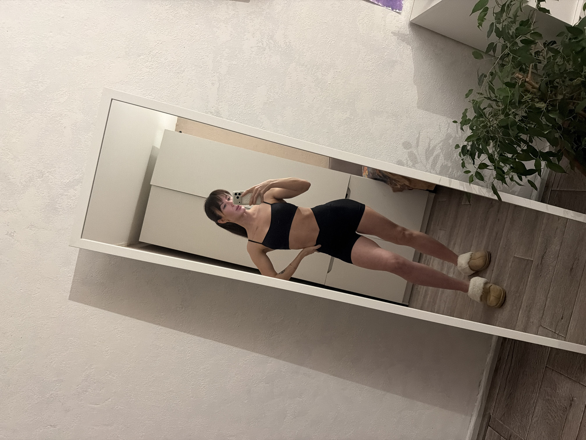 A young woman is captured in a mirror selfie within an indoor setting in Perm, Russia. The image is rotated diagonally, showing the woman standing in a large, white-framed mirror. She wears a black sports bra and black athletic shorts, complemented by fluffy light brown slippers. Her dark hair is styled with bangs, and she holds a white mobile phone in her right hand, aiming it towards her reflection to take the photo.
The background reflected in the mirror reveals a clean, modern aesthetic. To the left, a tall, white piece of furniture, possibly a cabinet or wardrobe, is visible. On the right, a leafy green houseplant, likely a ficus, adds a touch of nature to the room. The room's wall, partially visible outside the mirror frame and within its reflection, is painted a light color with a subtle textured finish. The floor, seen both inside and outside the mirror, features a light wood-grain pattern. The lighting appears to be artificial, suggesting the photo was taken indoors, possibly during the evening or night. The woman maintains a neutral expression, focused on her self-portrait. No visible text is present in the image.
