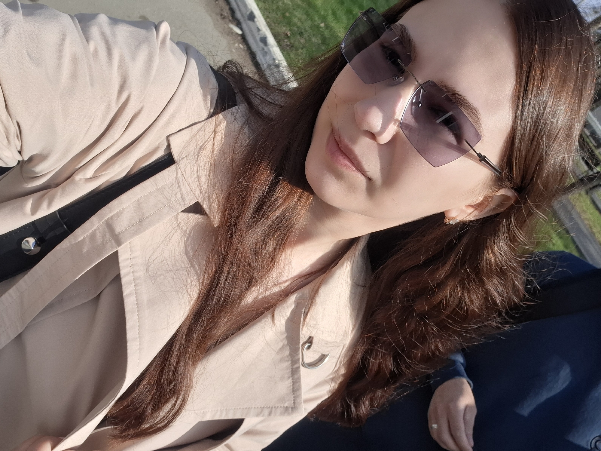 A woman is pictured outdoors in a sunny setting, likely in a park or green area. She has long, auburn hair and is wearing rimless sunglasses with a square frame, a light beige trench coat, and a light-colored top underneath. Her skin tone appears light to fair. A hand with a ring on the ring finger is visible in the bottom right corner, resting on a navy blue surface, possibly another person's clothing or a vehicle seat. A section of green grass and a paved area are visible in the background, suggesting an urban park setting. The picture has a casual, selfie style. The location is in Chaikovsky, Russia.