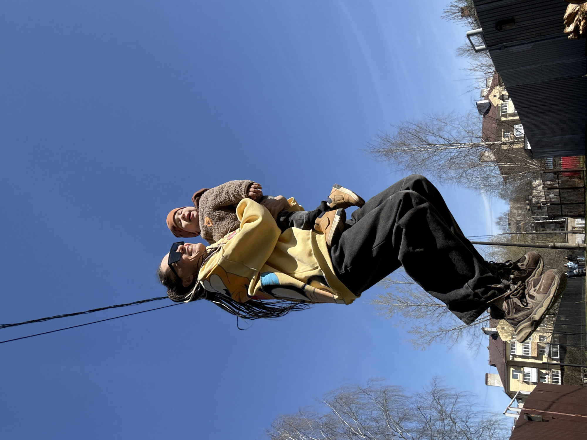 An outdoor, low-angle photograph captures an adult holding a young child against a clear, vibrant blue sky in Bor, Russia. The image is taken on a sunny day, likely in late winter or early spring, given the bare trees visible in the background.
The adult, positioned on the left side of the frame, is wearing a yellow hoodie with a colorful graphic design, dark trousers, and sturdy brown and black sneakers. They have long dark hair, possibly in braids or dreadlocks, and are wearing dark rectangular sunglasses. Their head is tilted back, and a wide smile is evident, conveying joy.
The young child is held by the adult, positioned slightly above and in front of them. The child is dressed in a textured, seemingly fuzzy, brown jacket or sweater and a matching brown beanie. The child also appears to be smiling, looking upwards with a happy expression. Their pose suggests they are being lifted or playfully swung, creating a dynamic and cheerful interaction.
The background, mostly visible on the right side of the frame, reveals an urban or suburban setting with several buildings featuring windows. Bare trees are interspersed among the structures, and a dark fence or wall runs along the far right edge. A single black wire or cable stretches horizontally across the upper part of the blue sky, creating a visual line. The bright sunlight and clear sky contribute to a lighthearted and joyful atmosphere. There is no visible text in the image.