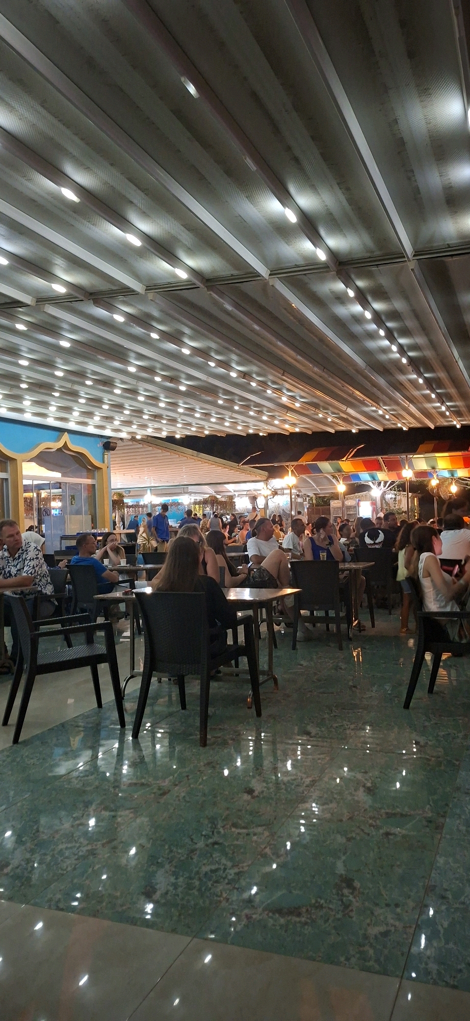 The image captures an outdoor dining area in Kemer, Turkey, under a retractable awning illuminated by strings of lights. The floor is a reflective, marbled green tile. A crowd of people are seated at tables, engaged in dining and conversation. In the foreground, black chairs and tables are visible. The background reveals more tables and patrons, with a colorful awning structure and architectural elements suggesting a commercial establishment. The lighting creates a warm, inviting atmosphere, typical of an evening at a restaurant or cafe.