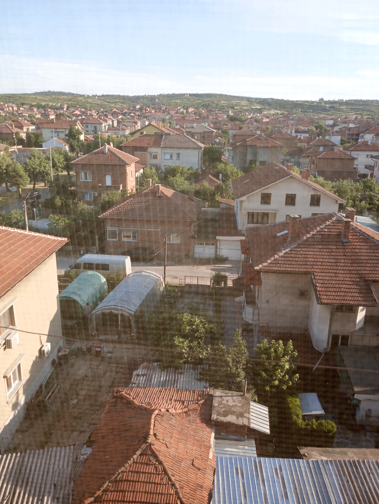 This is an aerial, eye-level photograph of a residential neighborhood in Harmanli, Bulgaria, bathed in the soft light of either early morning or late afternoon. The scene is characterized by a dense collection of red-tiled, single-family homes and smaller structures, tightly packed together. The architecture is predominantly traditional, with many buildings featuring exposed brick or light-colored facades.
In the foreground, the view is partially obstructed by a window screen, creating a subtle mesh overlay on the image. A light-colored, two-story building occupies the lower left quadrant, with an air conditioning unit visible on its exterior wall and a simple window on its upper floor. Adjacent to this building, and slightly further back, is a smaller building with a red-tiled roof.
Further into the scene, a paved area hosts two large, greenhouse-like structures covered in translucent plastic. One is green, the other is silvery-grey. A white van is parked near these structures. The surrounding yards and open spaces are a mix of dirt, gravel, and patches of greenery. A person, partially visible and dressed in darker clothing, appears to be inside or near one of the greenhouses.
Beyond these immediate structures, the landscape unfolds into a panorama of rooftops extending towards a tree-covered, rolling hill in the distance under a clear, pale blue sky. The overall impression is of a lived-in, established urban environment with a rustic feel.
