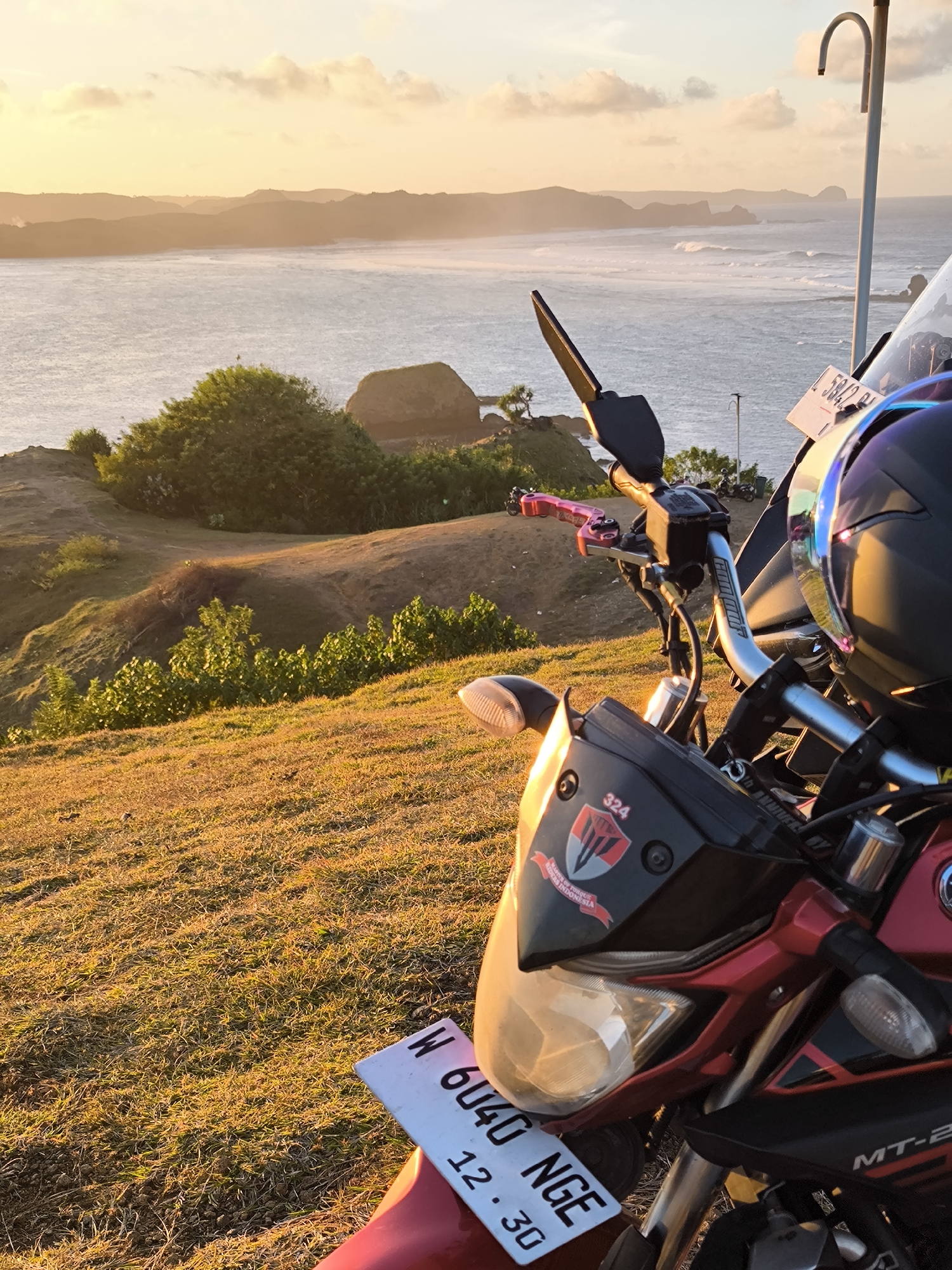 A medium, eye-level shot shows a red and black MT-25 motorcycle parked on a grassy knoll overlooking a large body of water, presumably the ocean. The foreground is dominated by the front of the motorcycle, with a prominent license plate reading "W 6040 NGE 12.30." A sticker with the number "324" and a stylized shield design is visible on the front fairing. The handlebars are fitted with pink levers. A dark helmet sits on the motorcycle seat, with a reflective visor catching the light.
In the midground, a green, grassy hill slopes down to the water's edge, dotted with bushes. A tan path cuts across the hill. In the background, the ocean stretches to the horizon, with several rocky landmasses visible in the distance. The sky is a mix of light yellow and orange, indicating either sunrise or sunset.
Based on the landscape and available information, this photo could be from Kuta, Indonesia. The hilly terrain and coastal views are consistent with that region. There are no people visible in the frame.