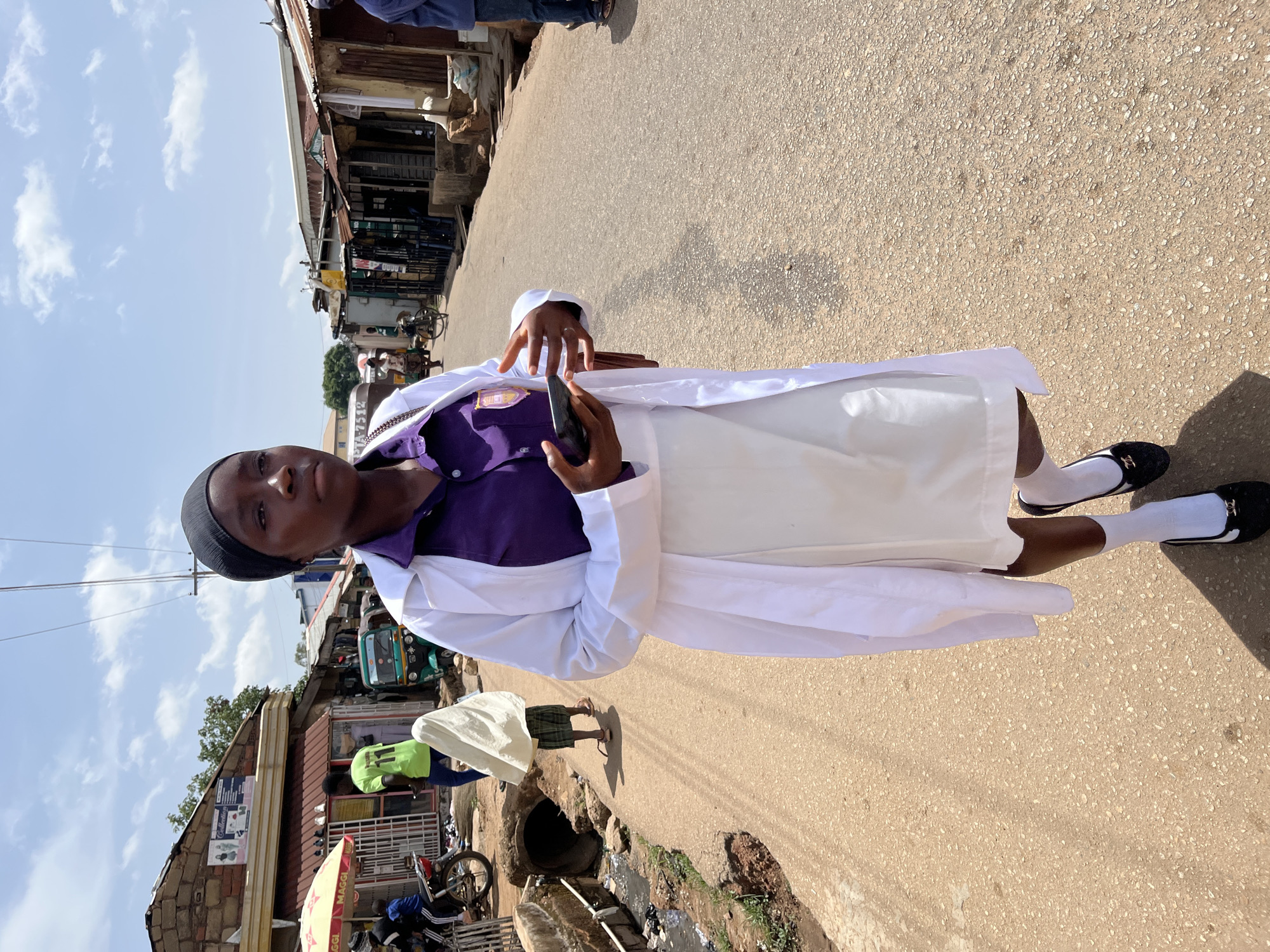 A young person in a white lab coat, purple shirt, white skirt, and black flats with white socks stands on a paved street in Gura Top, Nigeria. They hold a dark object in their hands. Buildings and a tricycle are visible in the background. The street is cracked and shows signs of wear. A sewer drain is partially visible at the bottom left. Other people are walking nearby. The sky is blue with scattered clouds.