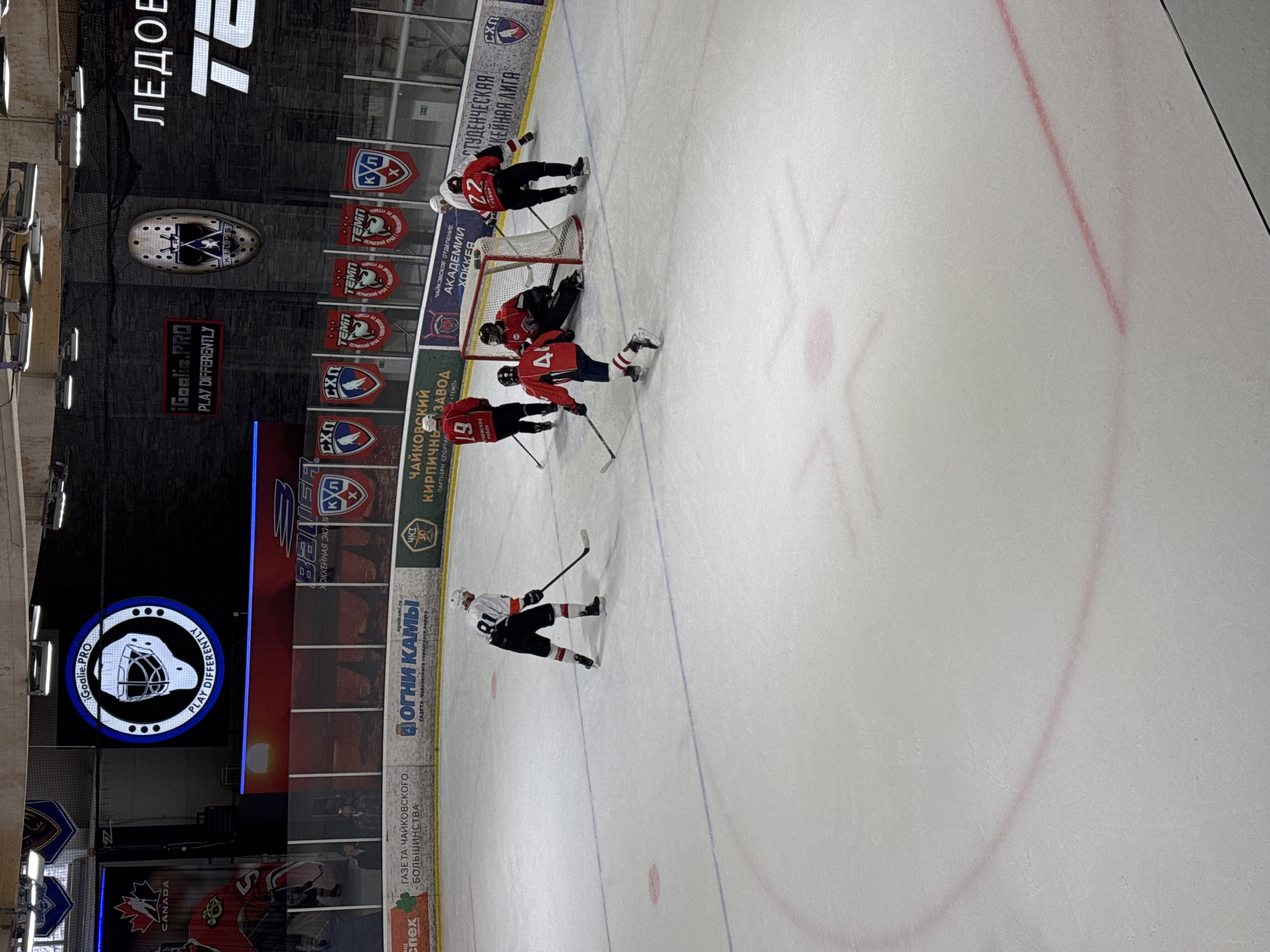 The image shows a hockey game in progress at an indoor arena in Chaikovsky, Russia. Several players in red jerseys are near the goal defended by a player in black. One player in a black jersey with number 80 on the back is positioned further away. The rink has red lines and circles painted on the ice. The background includes advertisements and logos of various organizations, including "Oгни Кам", "чайковский кирпичный завод" and "temo". A large digital display in the background features an image of a hockey mask with the logo "iGoalie.PRO PLAY DIFFERENTLY", and an arena scoreboard is also present.