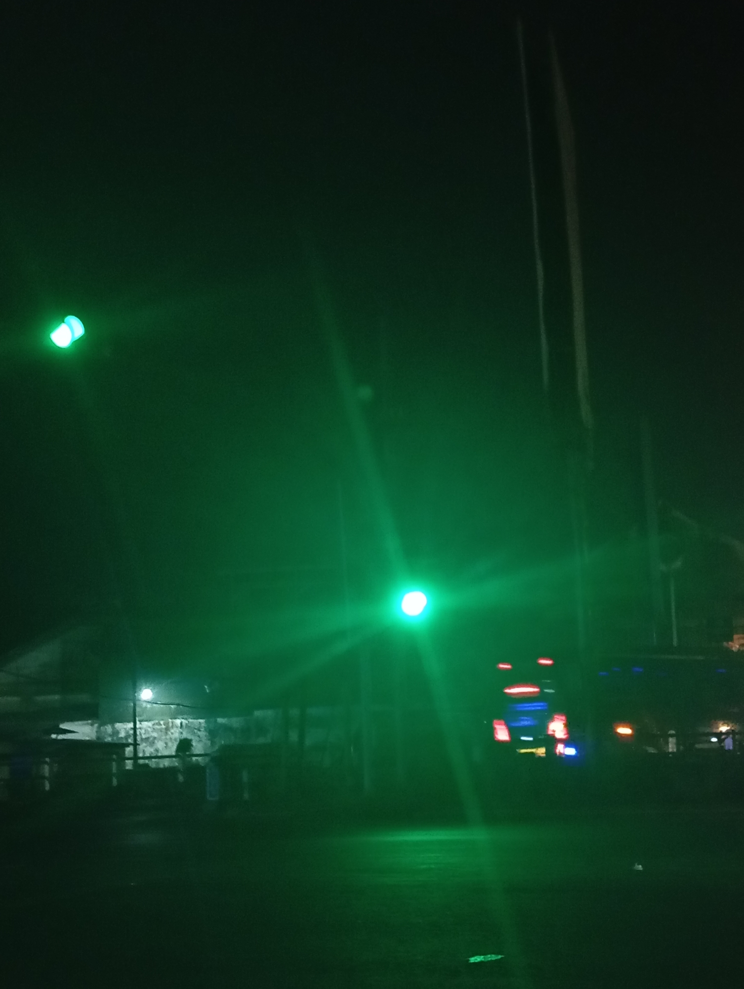 The image is a nighttime photograph taken in Sokaraja, Indonesia. It depicts a dark outdoor scene illuminated by various green lights and some red and blue lights from a vehicle. In the foreground, the ground appears to be a dark, possibly wet, surface. In the middle ground, there are structures that resemble outdoor seating or stalls, with some faint human figures visible. Further back, a vehicle with illuminated red and blue lights is present. Two prominent green lights are visible, one in the upper left and another in the middle of the frame, casting strong beams and creating a hazy effect in the dark atmosphere. Tall, thin vertical elements, possibly poles or wires, are also present in the background.