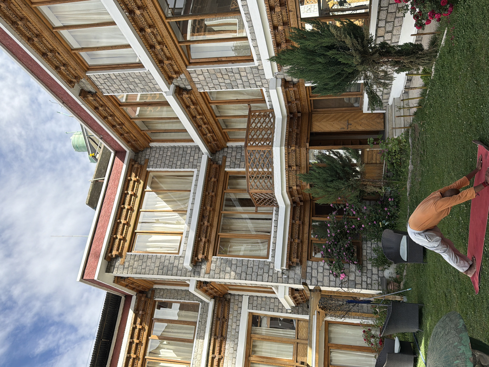 A person in an orange shirt and grey pants performs a yoga pose on a red mat in a grassy courtyard in front of a multi-story building in Leh, India. The building is constructed of stone and wood, with ornately carved wooden trim around the windows and roofline. There are several windows with white curtains and a wooden balcony. To the left of the building are two dark wicker chairs, and a pink flowering bush. To the right of the building are two evergreen trees and a cluster of pink roses. The sky above is cloudy.