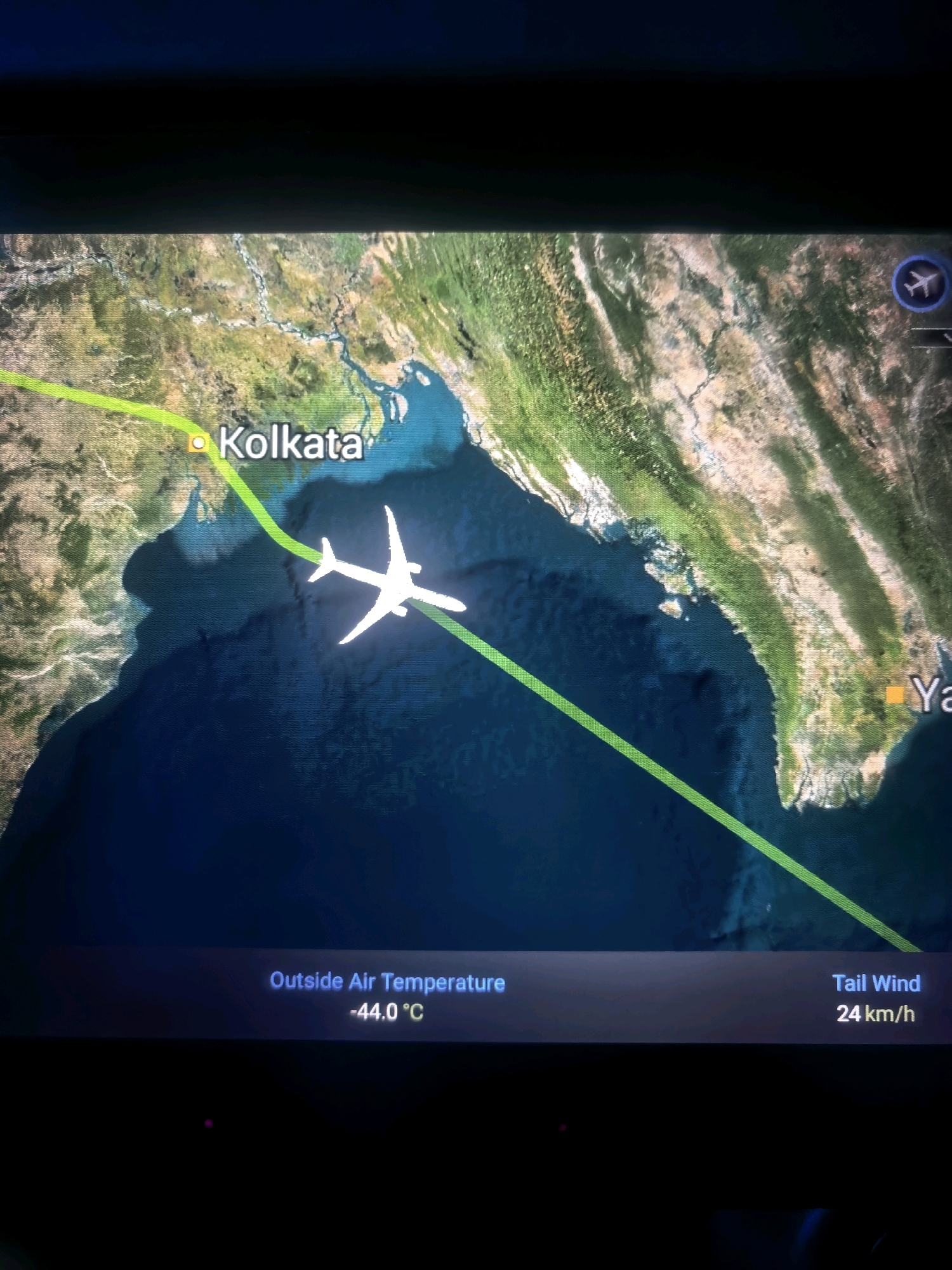 The image displays a flight path on a map displayed on a screen. The flight path, marked by a green line, originates near Kolkata, India, and extends towards a destination labeled "Ya" which is likely Yangon, Myanmar. A white airplane icon is shown on the flight path. The map shows a portion of the Indian subcontinent and the Bay of Bengal. The screen also displays flight information: "Outside Air Temperature -44.0 °C" and "Tail Wind 24 km/h". The city of Singapore is not visible or mentioned in the image.