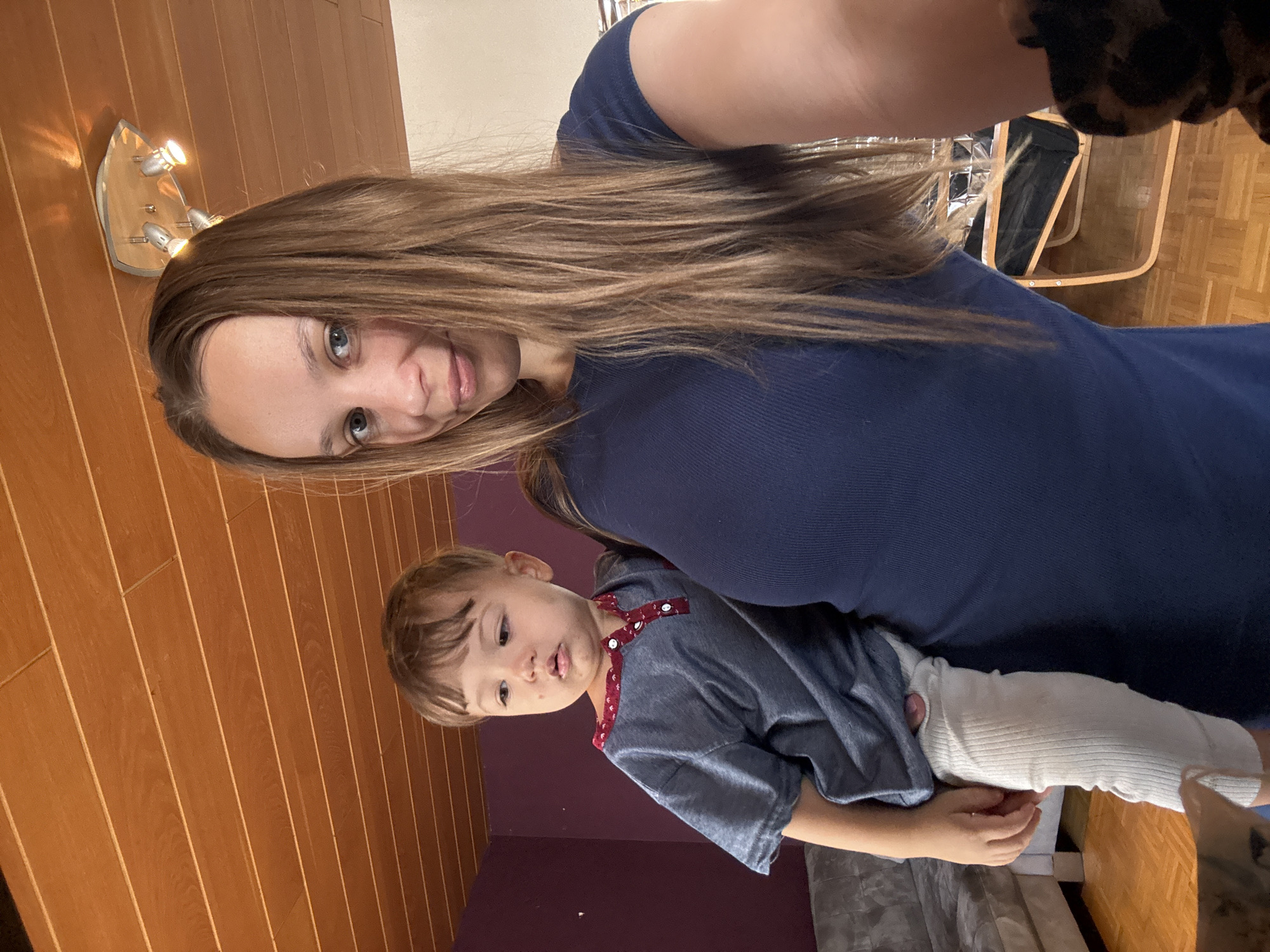 A young woman with long, brown hair, wearing a navy blue t-shirt, holds a toddler in her left arm. The toddler, wearing a grey shirt and white pants, looks away from the camera. The scene appears to be indoors, possibly in a residential setting, with wooden-paneled walls and ceiling visible. The lighting suggests daytime. There is no indication of the location being in Überherrn, Germany.