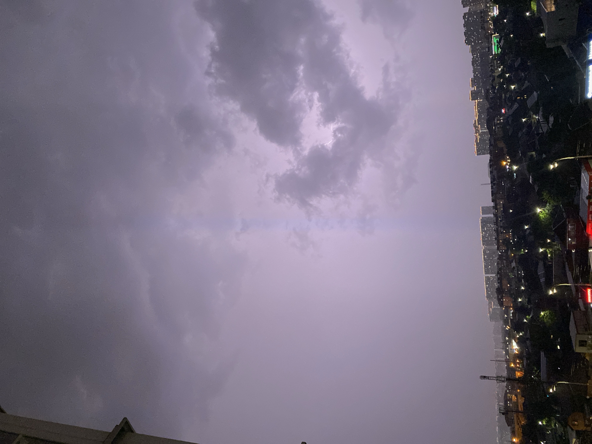 A cityscape at night is depicted, with a dramatic sky filled with dark, stormy clouds. A bright bolt of lightning splits the sky vertically, illuminating the clouds with a faint purple glow. The city below is dotted with lights from buildings and streetlamps, with rows of residential buildings and some industrial structures visible. The overall impression is one of a powerful natural event occurring over an urban environment.