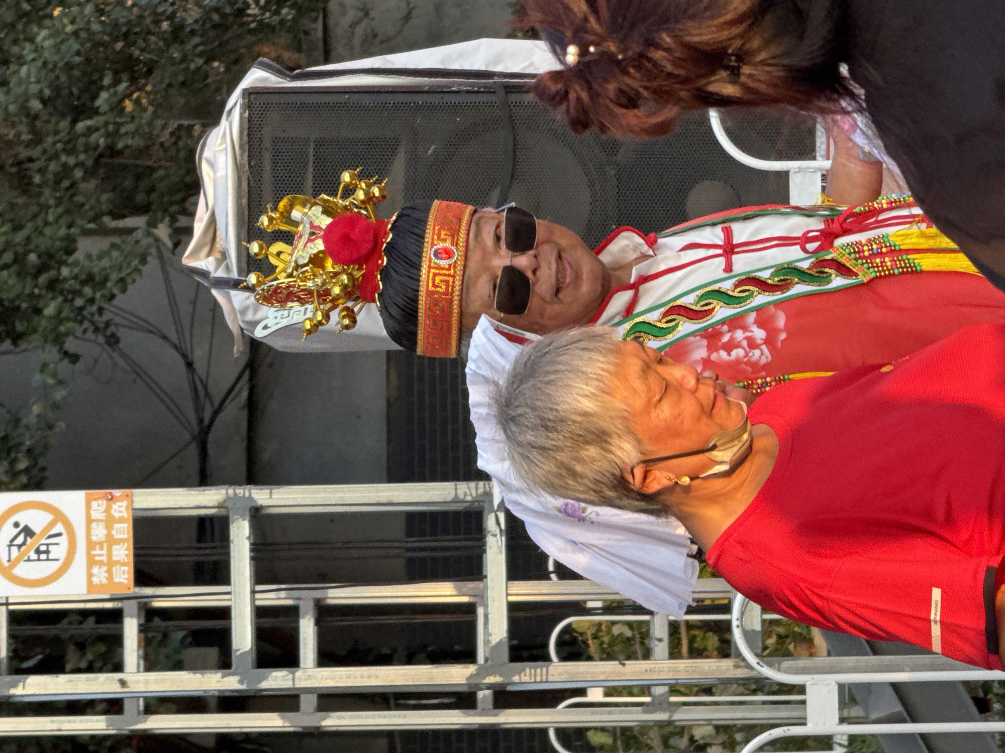 The image depicts an outdoor scene in Jinzhou, China, likely during a festive or cultural event. In the foreground, two individuals are partially visible. On the left, a woman with short gray hair wears a vibrant red shirt and a face mask hanging around her neck. To her right, a man is dressed in traditional attire, including an ornate headdress with gold embellishments and red accents, sunglasses, and a white top with red and green trim. He appears to be the central focus, smiling towards the viewer.
Behind the individuals, a large speaker is partially visible, draped with a light-colored fabric. To the left of the scene, a metal ladder structure is present, with a sign attached to it bearing Chinese characters and a prohibition symbol. The background consists of trees and some building structures, suggesting an urban environment. The lighting indicates it is daytime, possibly late afternoon due to the warm tones. The overall atmosphere seems to be one of a public gathering or celebration.