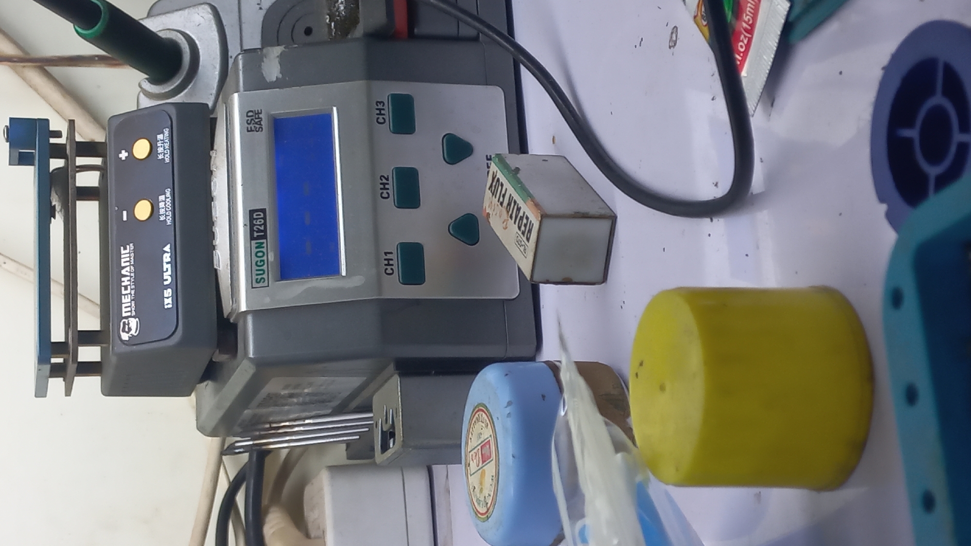 The image depicts a workbench cluttered with electronic repair equipment. In the foreground, a bright yellow cylindrical cap is prominently displayed, with a blue and white container to its left and a small, rectangular box labeled "REPAIR FLUX" to its right. A thin, translucent object, possibly a plastic tube, lies diagonally across the yellow cap.
Dominating the mid-ground is a silver and gray electronic device with a blue digital display labeled "SUGON T26D". Below the display are three green buttons labeled "CH1", "CH2", and "CH3", and two smaller green triangular buttons. To the left of this device is a black metal stand holding several thin metallic rods.
In the upper portion of the image, a dark gray unit with a blue top plate is visible, featuring branding that includes "MECHANIC" and "IX5 ULTRA". This unit also has yellow buttons with Chinese characters and English text like "HOLD COOLING" and "HOLD HEATING". A green-tipped tool, possibly a soldering iron, is attached to the right side of the workbench.
The background reveals more of the workspace, with visible wires, power outlets, and portions of other equipment. The overall environment suggests an active electronics repair station. The specific location within Jetis Indah, Indonesia, cannot be determined from the visual information provided in the image. There are no people or discernable actions taking place; the scene is static and focused on the arrangement of tools and equipment.