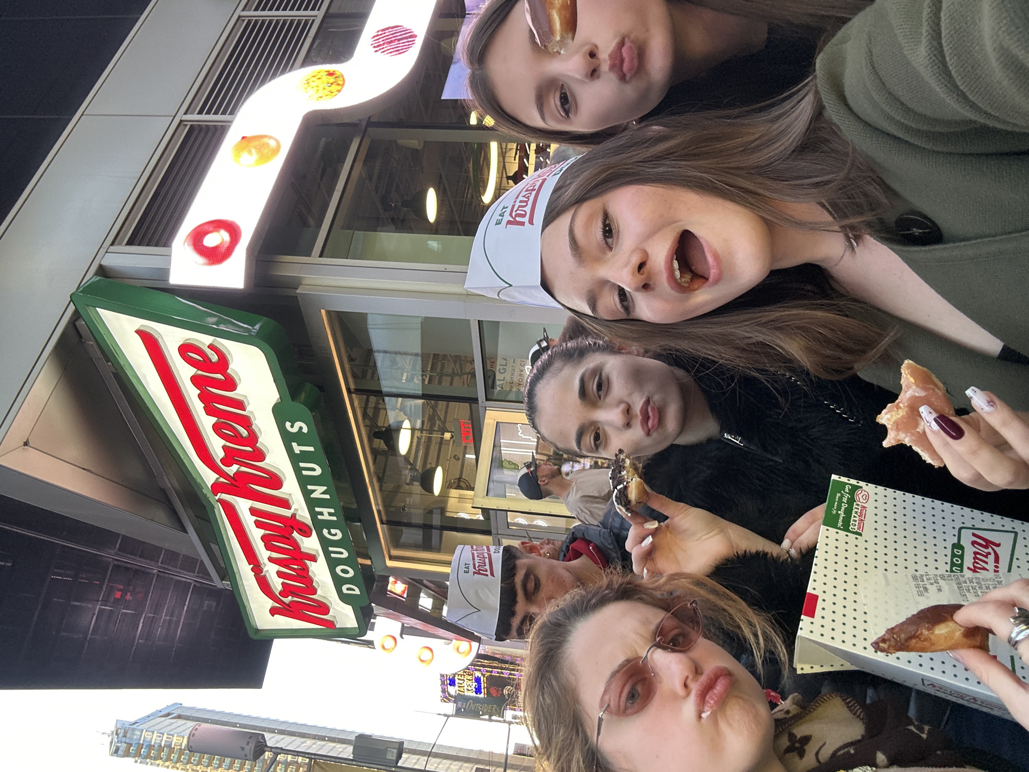 The image captures a group of five young women posing for a selfie outside a Krispy Kreme Doughnuts store. The store's prominent green and red logo sign is visible, along with a sign featuring illuminated donut shapes. The women are holding and eating donuts, with one woman wearing a Krispy Kreme employee hat. A Krispy Kreme box is visible, displaying text related to free doughnuts and a phone number. The surroundings include a building façade, and in the background, there is an illuminated sign indicating "YORK CI" and "AL GLA". The scene is in New York City, United States of America.