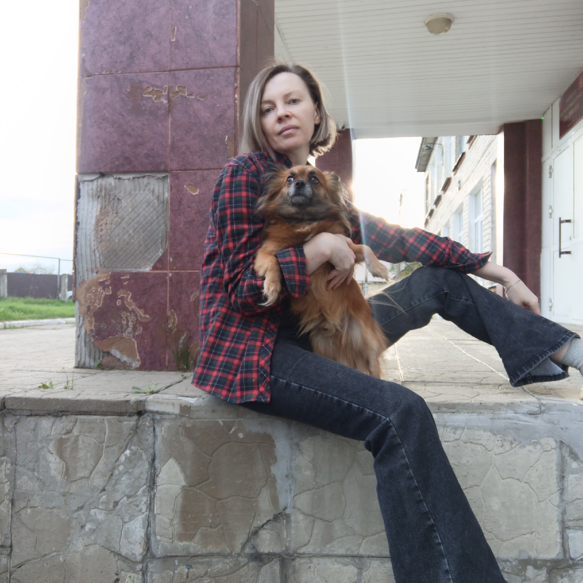 A woman sits outside a building in Chaikovsky, Russia, holding a small brown dog. The woman has fair skin and short blonde hair and is wearing a red and black plaid shirt and dark denim jeans. She is seated on a concrete wall, with a building behind her. The building appears to be constructed of red and white bricks. There are small patches of grass and vegetation visible in the foreground.