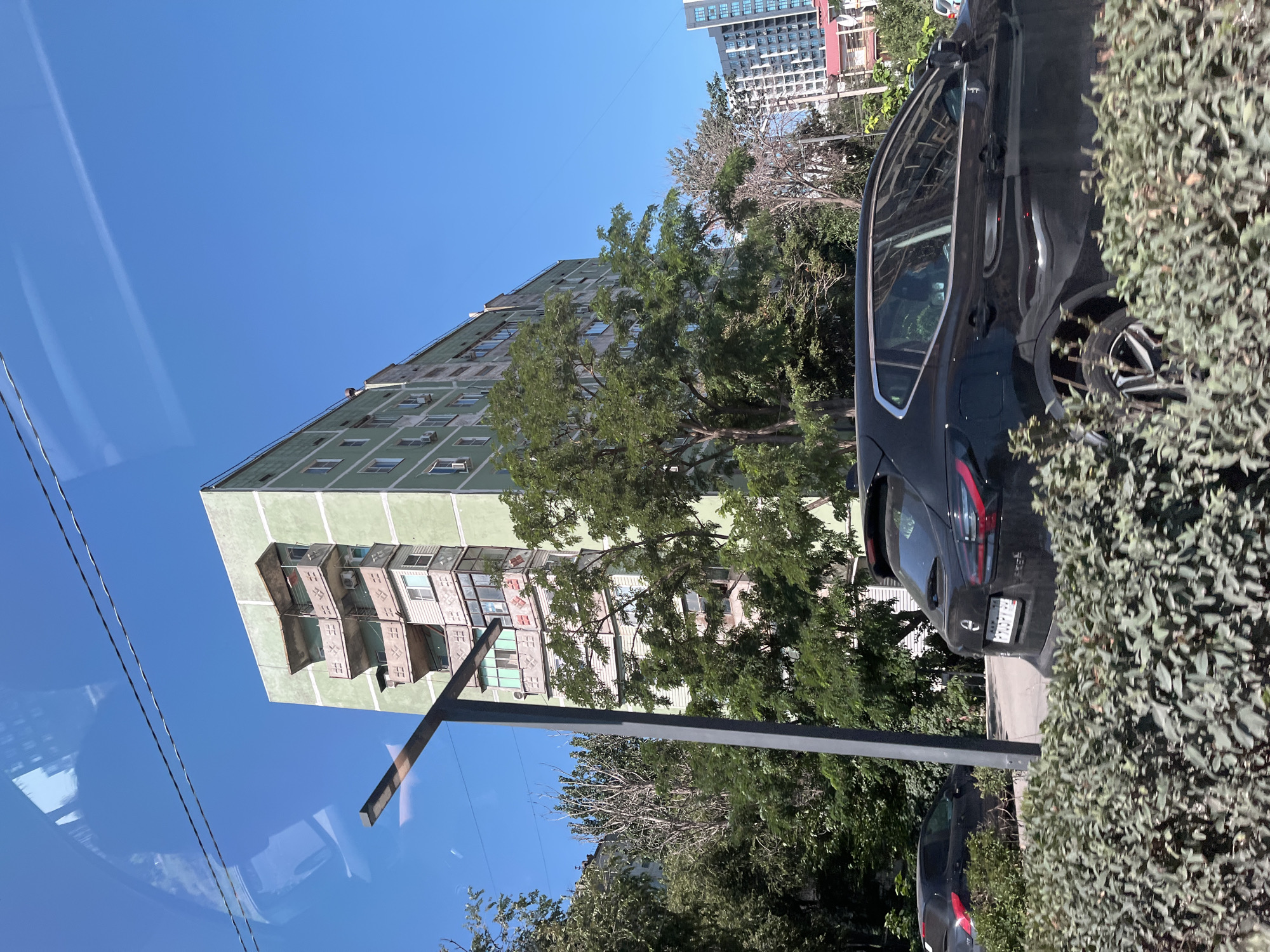 In Astrakhan, Russia, a black Nissan Qashqai is parked in front of a light green, multi-story apartment building under a clear blue sky. The building features numerous balconies with decorative railings, some of which appear to be enclosed. Trees with lush green foliage surround the building and line the street. A dark-colored sedan is visible parked further down the street. A modern street lamp stands on the left side of the image. In the background, taller modern buildings can be seen. The image appears to have been taken from a moving vehicle, as evidenced by the reflections on the left side of the frame and the slightly blurred foreground foliage.