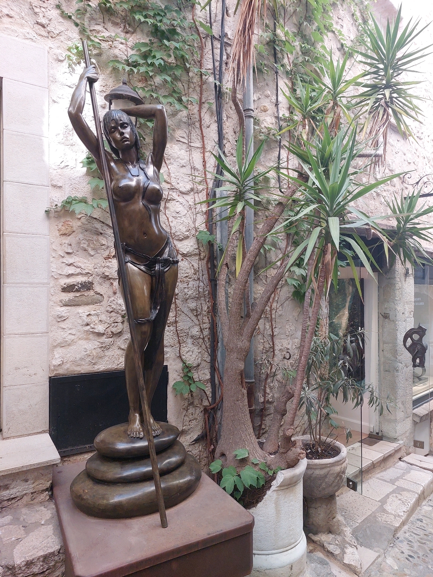 The image shows a bronze statue of a nude woman holding a long pole, placed in an outdoor courtyard. The statue stands on a stack of three smooth, dark-colored, rounded stones. The woman is depicted with her arms raised, one hand gripping the pole, and her head tilted back slightly. She wears a draped cloth around her waist. The background features a rough, stone-built wall partially covered with ivy. Several plants, including yucca and a dry, leafless tree with rough bark, are situated around the statue. To the right, there is a glass door leading into a building, with stone steps and a cobblestone pathway visible. The setting appears to be a narrow, somewhat shaded alleyway within a historic town. This scene is located in the city of Saint-Paul-de-Vence, France, known for its art galleries and charming medieval architecture.