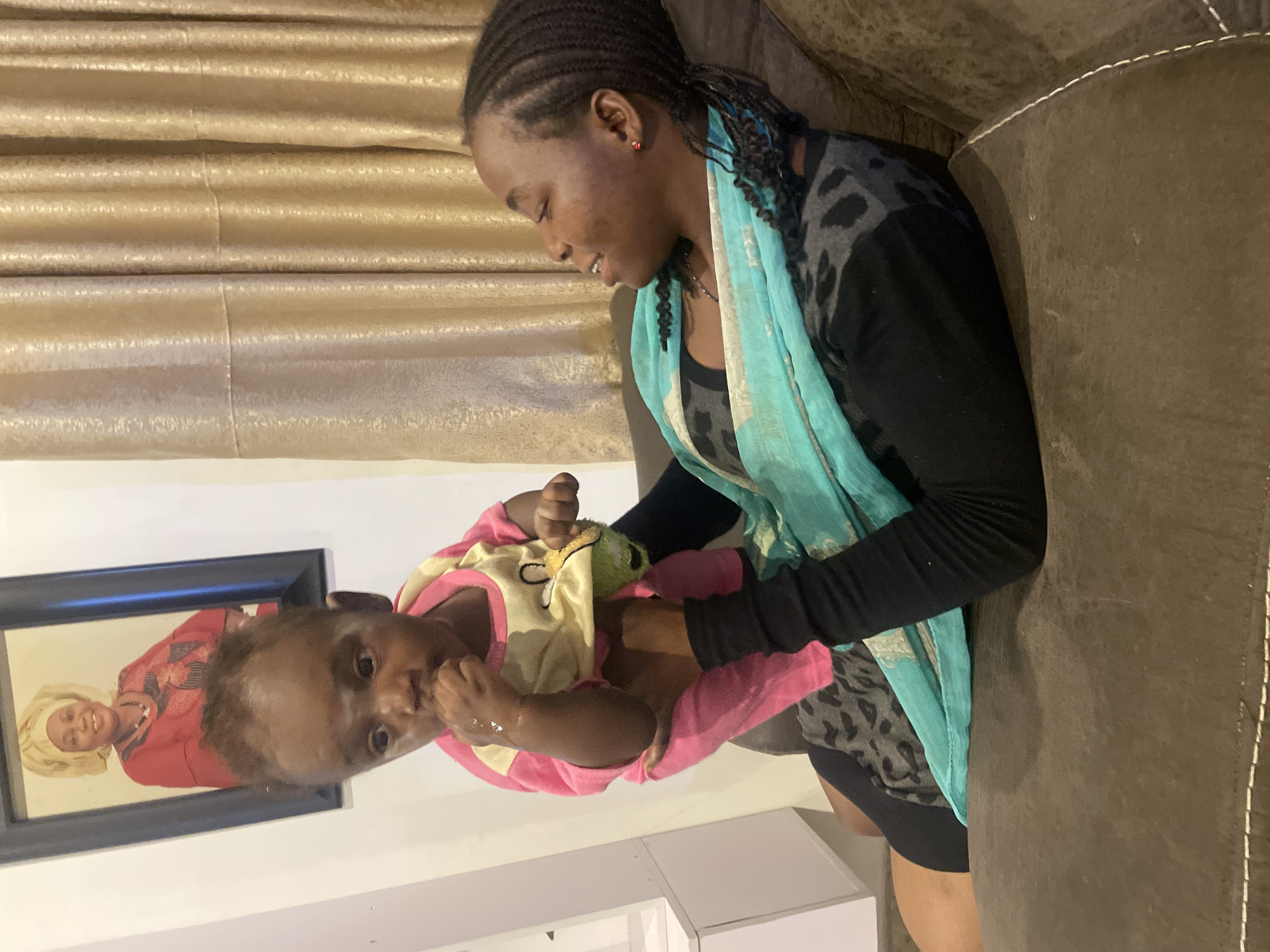 A woman and a baby are indoors. The woman, wearing a black long-sleeve shirt with a gray patterned undershirt, a turquoise scarf, and braids, is sitting on a brown couch. She is looking down at the baby who is wearing a pink and yellow onesie and holding a green object. A framed photo hangs on the white wall behind them. The location is likely in a home in Jos, Nigeria.