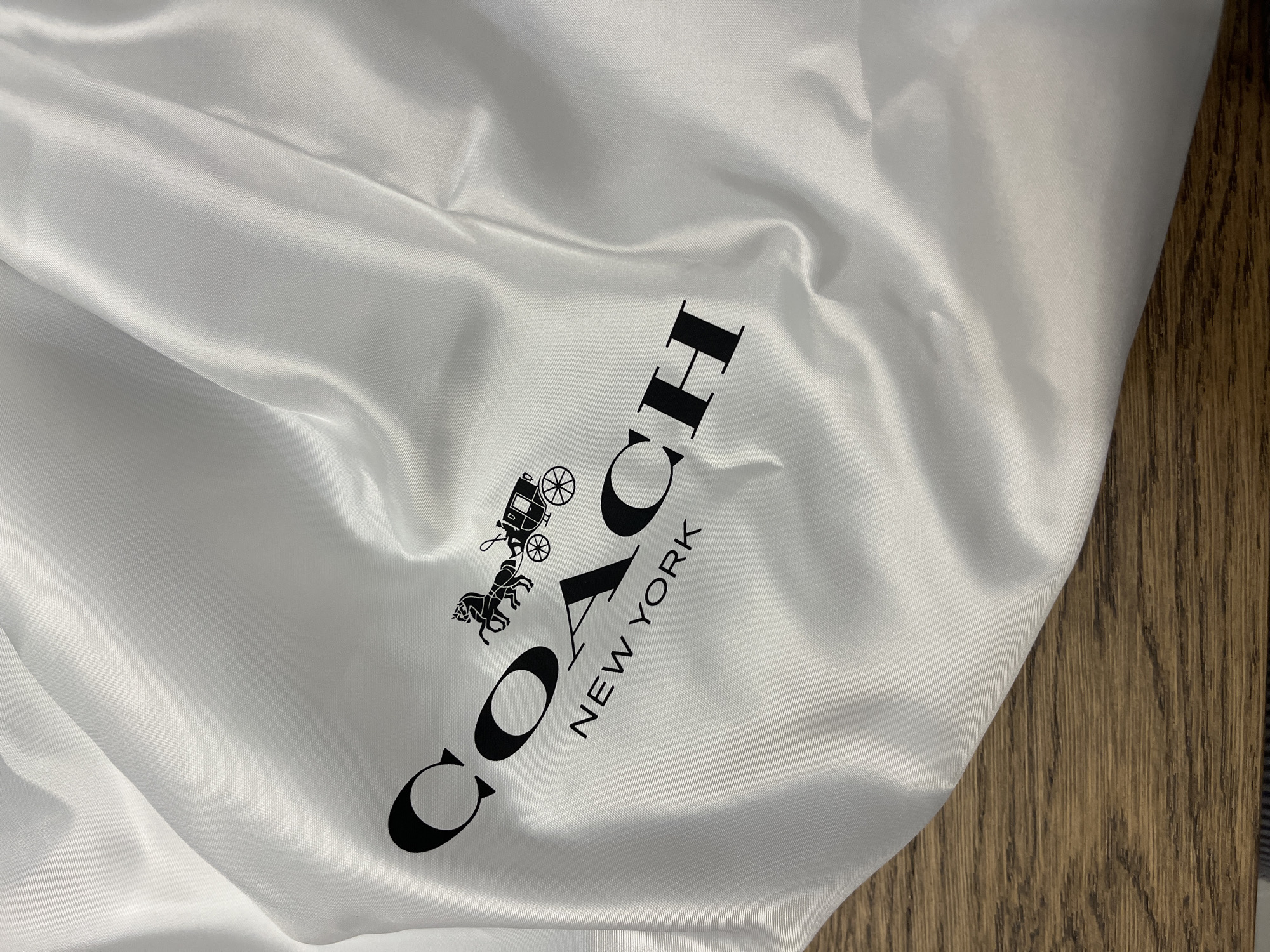 The image shows a close-up of a white fabric dust bag with the Coach logo and "NEW YORK" printed in black. The fabric is draped and creased, suggesting it is being held or has been folded. The texture of the fabric is visible, with a subtle sheen. A wooden surface is visible in the lower part of the image, suggesting the dust bag is resting on a table or floor. There are no people or animals present. The image does not provide any information about the city of Limassol, Cyprus, or any specific location within it.
