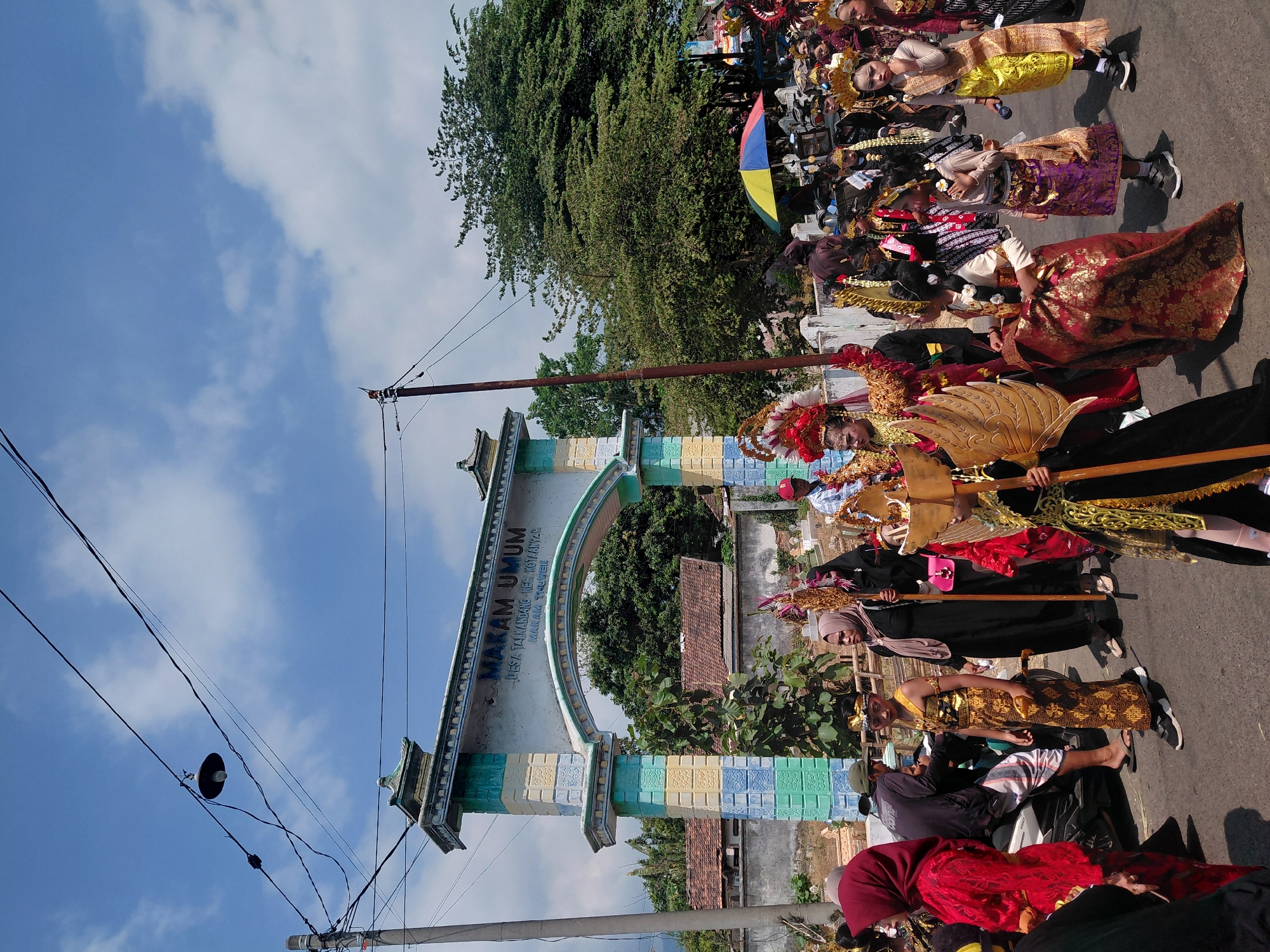 This image captures a street scene in Bimo, Indonesia, during a parade or cultural event. A decorated archway stands prominently, bearing the inscription "MAKAM UMUM" (Public Cemetery) and "DESA TALIKANDANG KEC. KOTANINYAR." The archway is adorned with colorful tiles in blue, green, and yellow.
A procession of people, many in traditional costumes, is walking along the road. Children are dressed in ornate attire, including headdresses and colorful fabrics. Some are carrying elaborate golden props. Adults are also present, some in traditional religious attire like hijabs. A few people are seated on motorcycles, and an umbrella is visible in the background.
The environment appears to be a rural or semi-urban area, with houses with tiled roofs and lush green trees. The sky is bright blue with scattered clouds. The overall impression is that of a lively community event taking place on a sunny day.