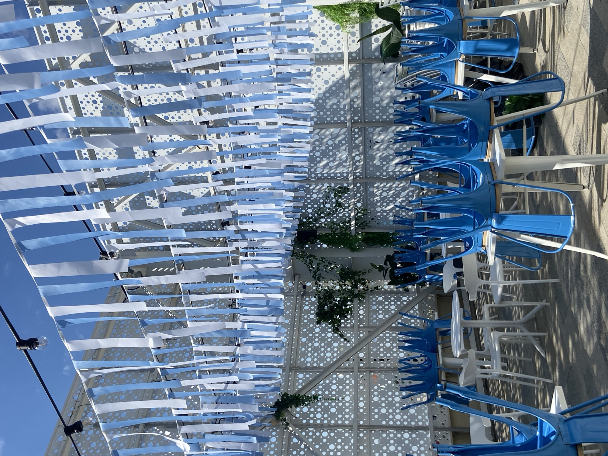 The image shows an outdoor seating area with a modern, geometric design. Above the seating area, there's a decorative installation of alternating blue and white strips of material, resembling a festive garland. The structure supporting the strips is white with circular cutouts.
To the right of the strips, there are stacks of blue metal chairs alternating with white tables. There are some leafy green plants integrated into the structure. The background is a concrete or paved area. A small part of a blue sky is visible in the upper left.
I'm unable to confirm the location in the city of Saratov, Russia.