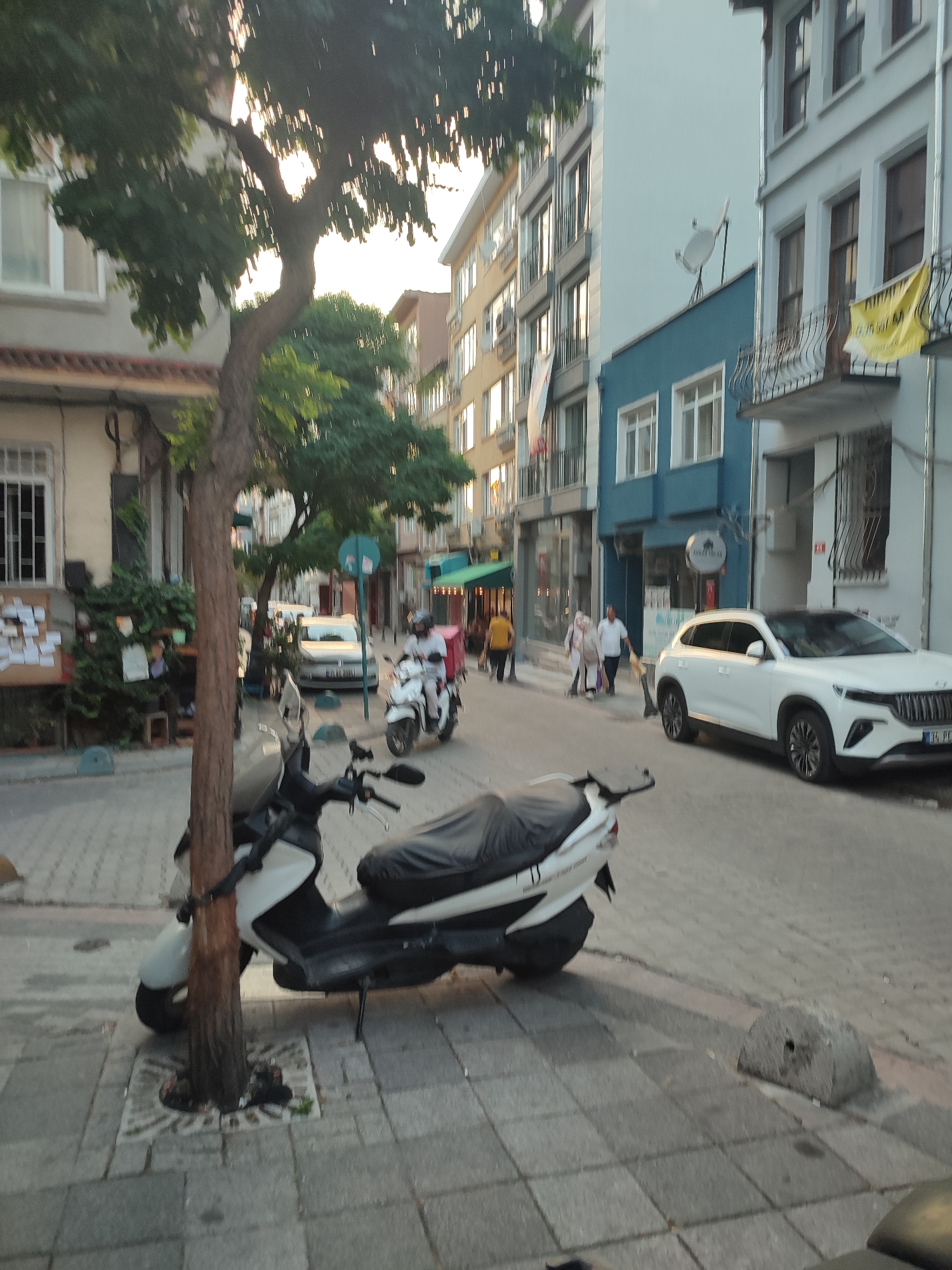 The image depicts a street scene in Kadıköy, Turkey. A tree grows on a sidewalk, with a parked scooter in the foreground. A white SUV is parked on the street, with a building and other vehicles in the background. Several people are walking on the sidewalk. The sky is overcast, and the lighting suggests it is late afternoon or early evening.
