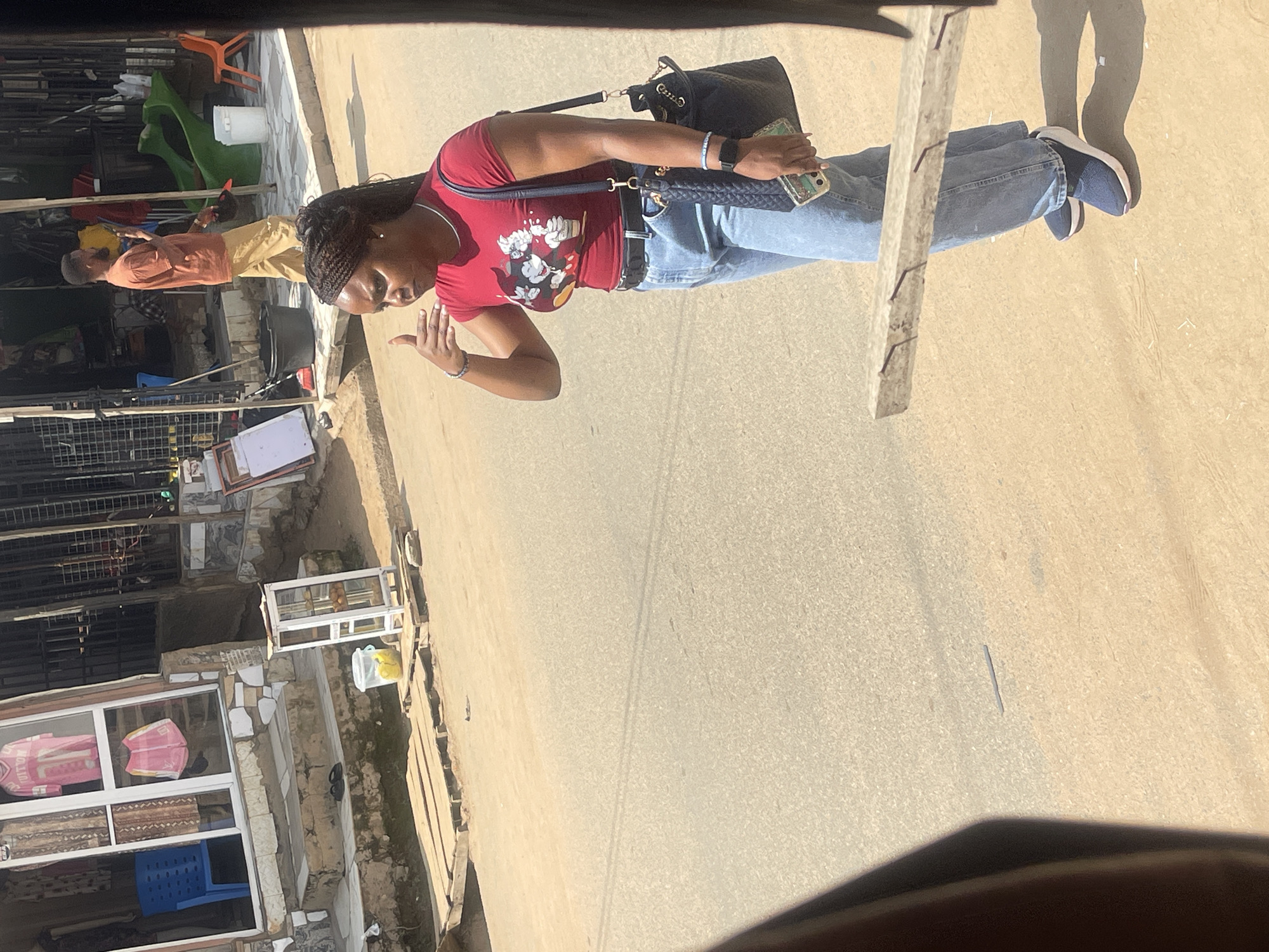 The image shows a young Black woman standing on a paved street in what appears to be an outdoor market or commercial area in Abuja, Nigeria. She is wearing a red t-shirt with a graphic print, blue jeans, and dark sneakers. A dark-colored handbag is slung over her right shoulder, and she is holding a smartphone in her right hand. Her left hand is raised to her ear, in a gesture that could indicate she is on the phone or signaling.
In the background, there are several shops with open fronts, displaying various goods. One shop has a blue chair visible through its window, and clothing items are displayed on shelves. Further back, a man in an orange shirt and tan pants is walking away from the camera, holding a dark object that might be a hat. There are also some children's playground equipment, including a green slide, and various other objects typical of a street market environment, such as buckets and a display case. The ground is dusty and uneven, with some wooden planks laid out on the road. The lighting suggests it is a bright, sunny day. The overall scene depicts a bustling, everyday urban setting in Abuja.