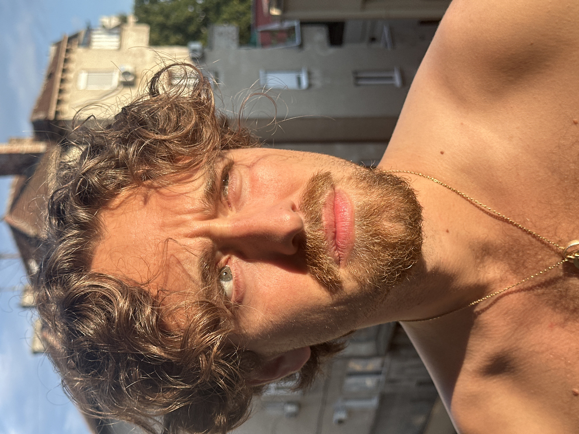 A close-up portrait shows a young man with curly brown hair and a goatee against a blurred urban background under a clear sky. The man's face is illuminated by the warm glow of what appears to be a sunset. His eyes are a light blue-green, and he is looking off to the right of the frame with a slight frown. He is wearing a thin gold chain around his neck. The background consists of buildings, one of which has a red-tiled roof and several windows, suggesting an urban setting, possibly Belgrade, Serbia, given the architectural style and common building materials observed. The lighting creates strong shadows and highlights on the man's skin, indicating the sun is low in the sky.