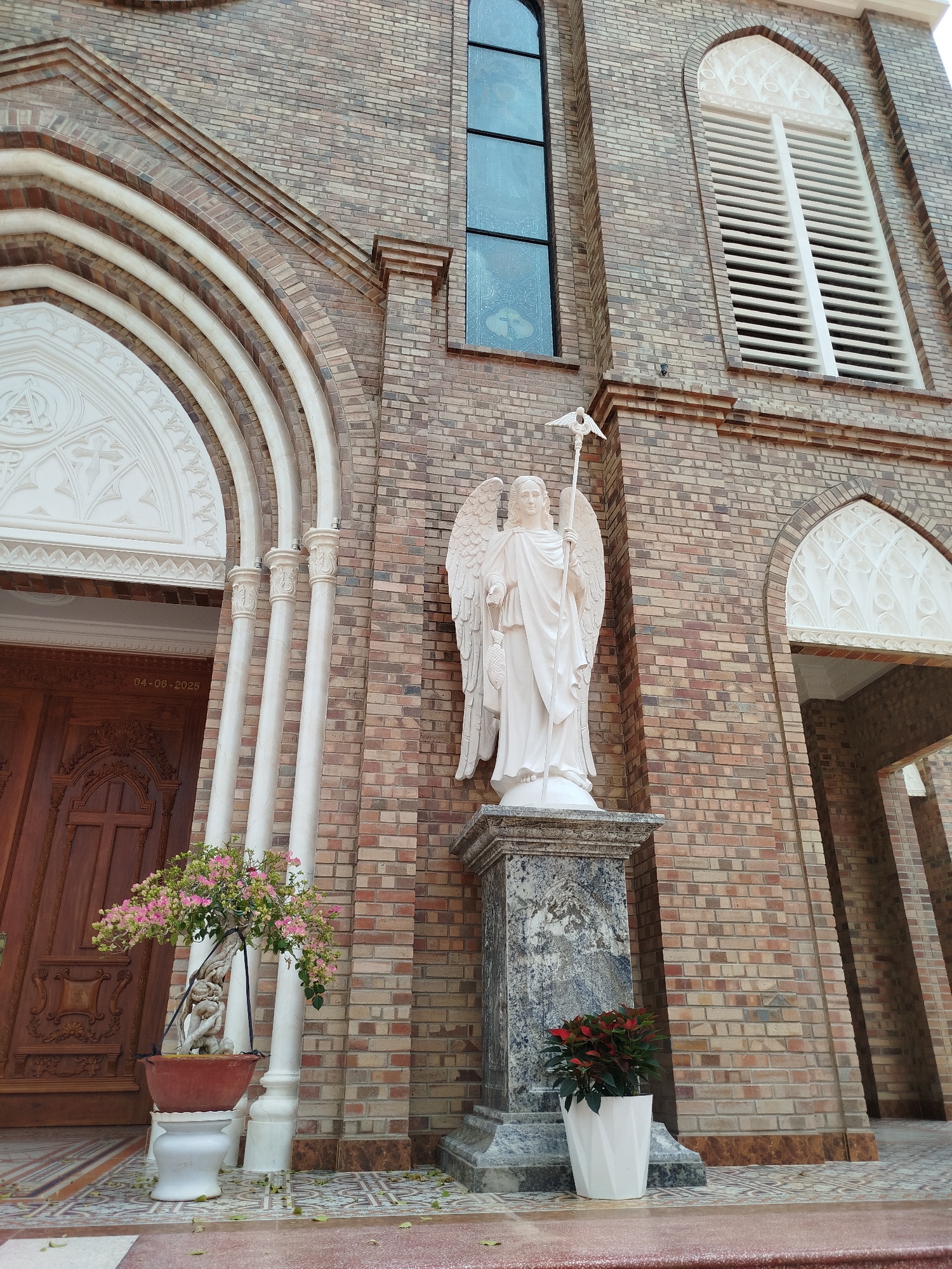 The image shows a street-level view of a brick building, which is the Cam Ly Church in Đà Lạt, Vietnam. The architecture features arched doorways with white trim, a window with stained glass, and a louvered window. A white angel statue stands on a marble pedestal in front of the building. A potted plant with pink flowers sits on a white base to the left of the statue. Another potted plant with red leaves is positioned to the right of the statue in a white pot. The foreground shows a patterned tiled floor.